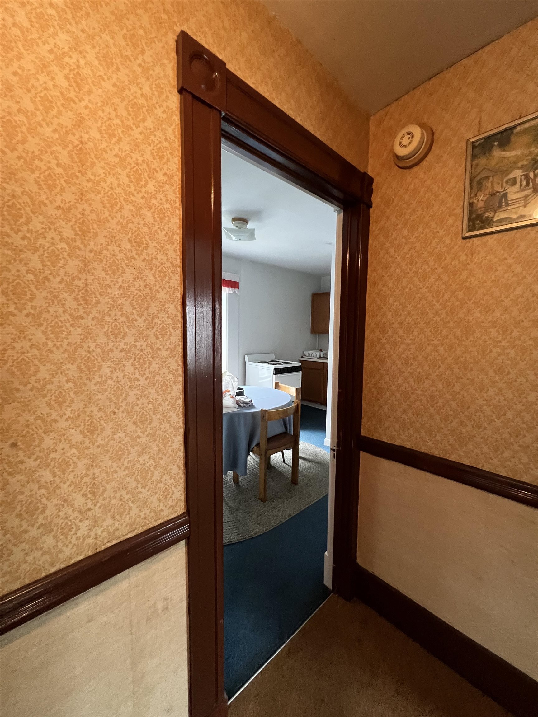 Kitchen and dining area seen through a dark wood-trimmed doorway from a patterned hallway.