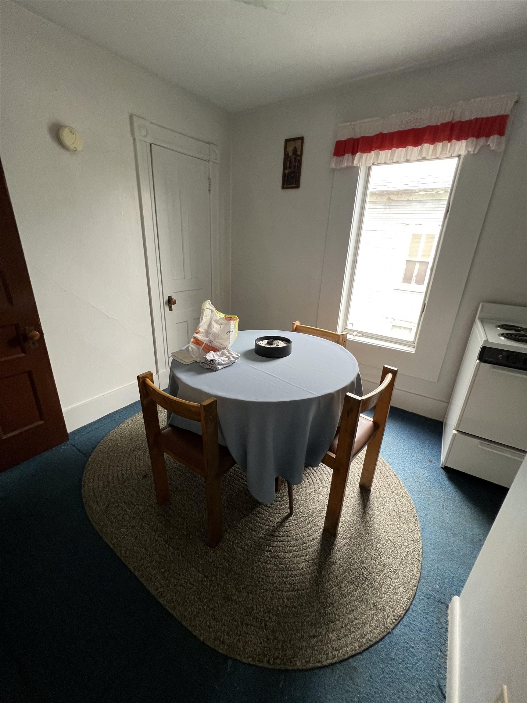 Small dining area with a round table, wooden chairs, woven rug, and red-and-white window curtains.