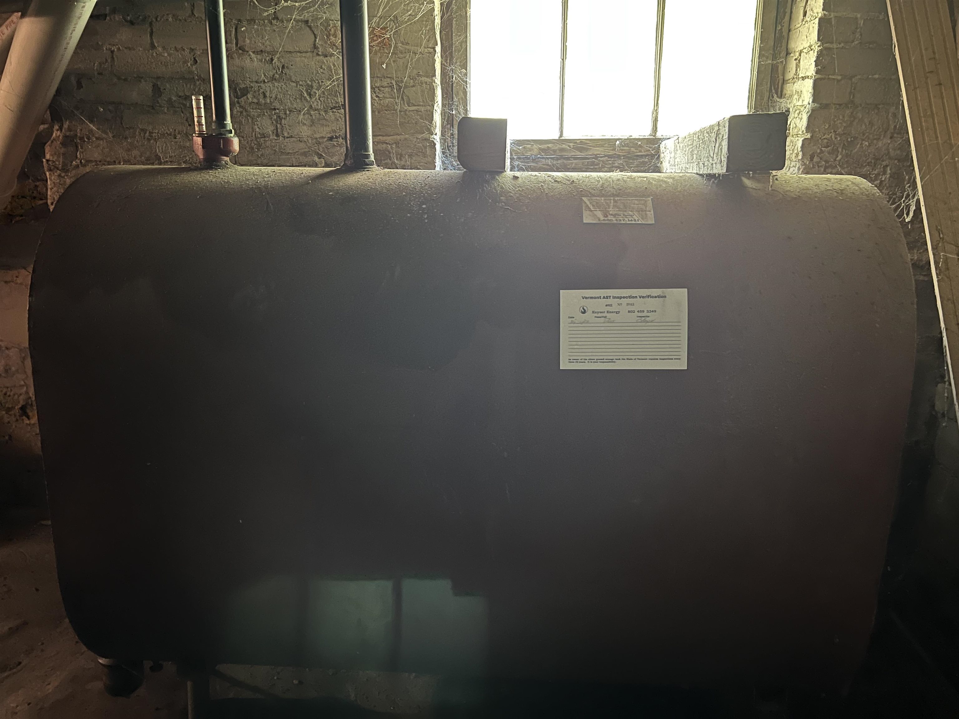 Basement heating oil tank beneath a window, with pipes and exposed stone foundation.