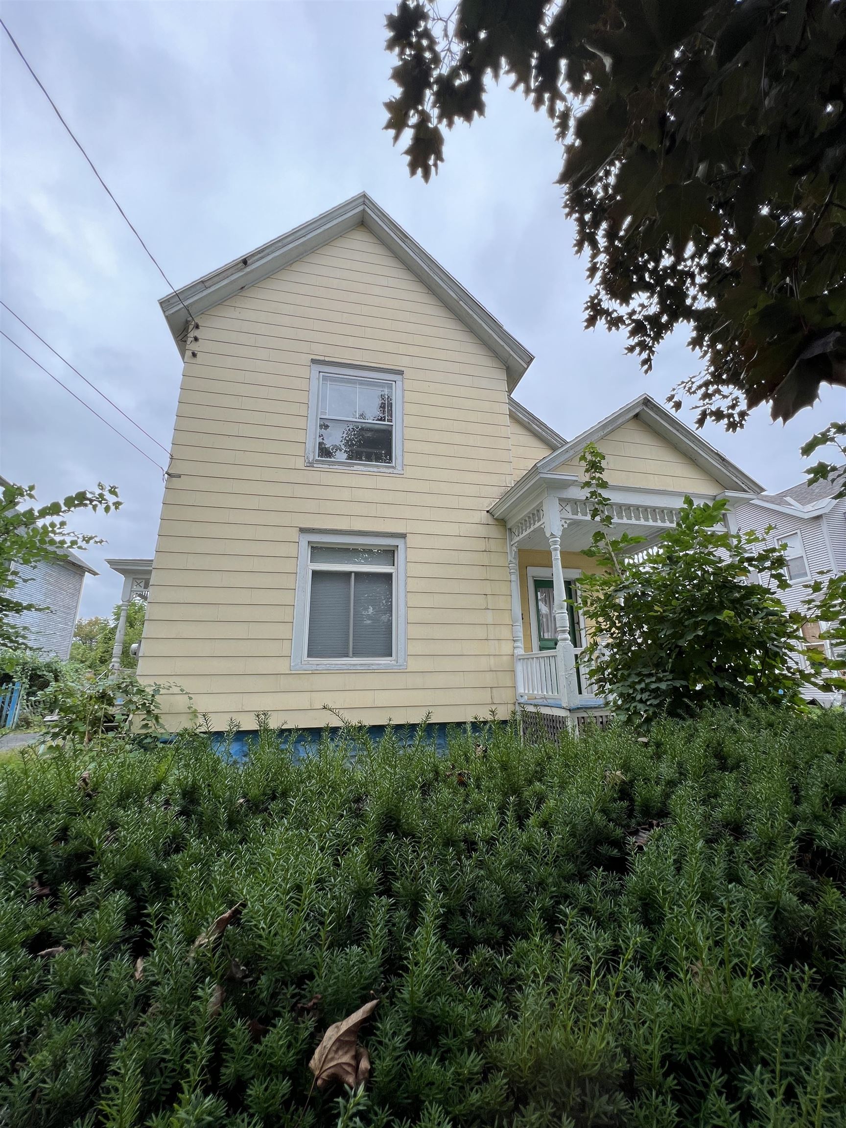 Pale yellow two-story house with white trim and a covered porch, partly obscured by dense shrubs.