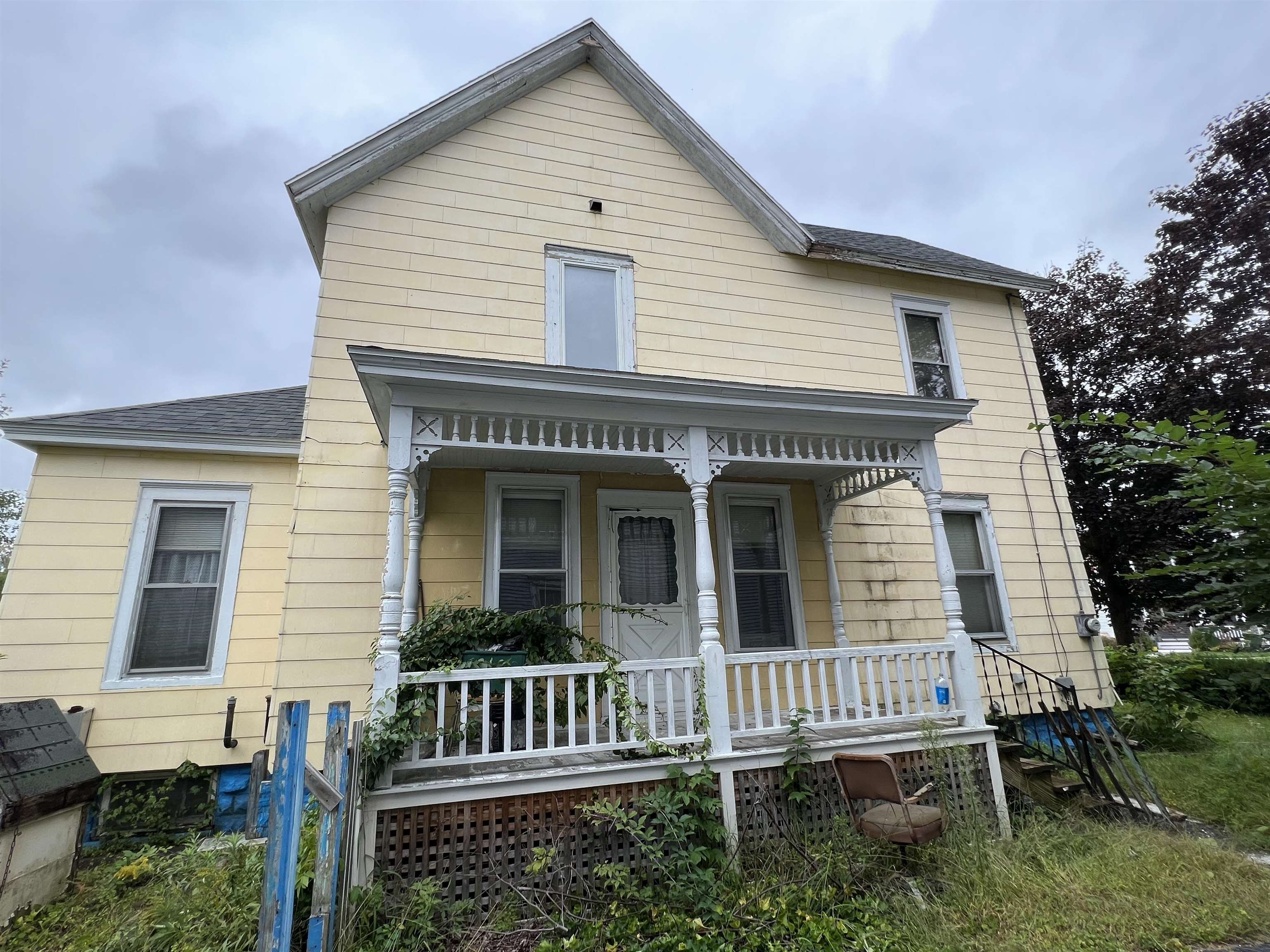 Two-story yellow house with a white decorative porch and overgrown plants in the yard.