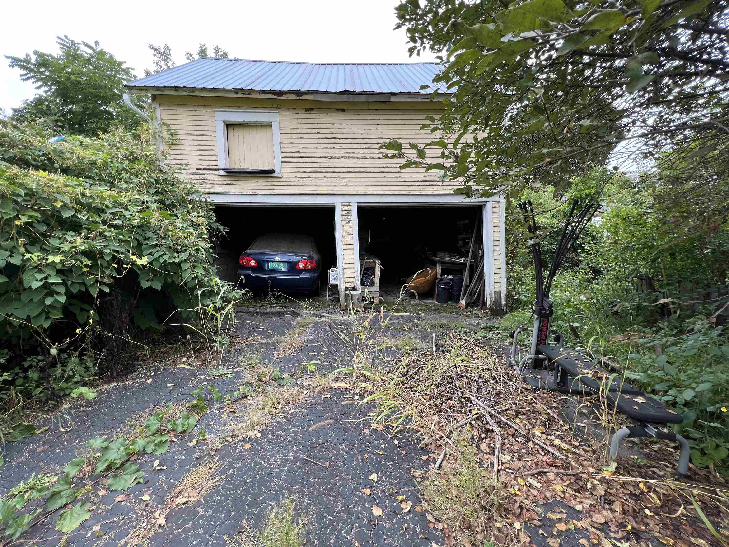Weathered two-bay garage with a car in one open bay and an overgrown driveway.