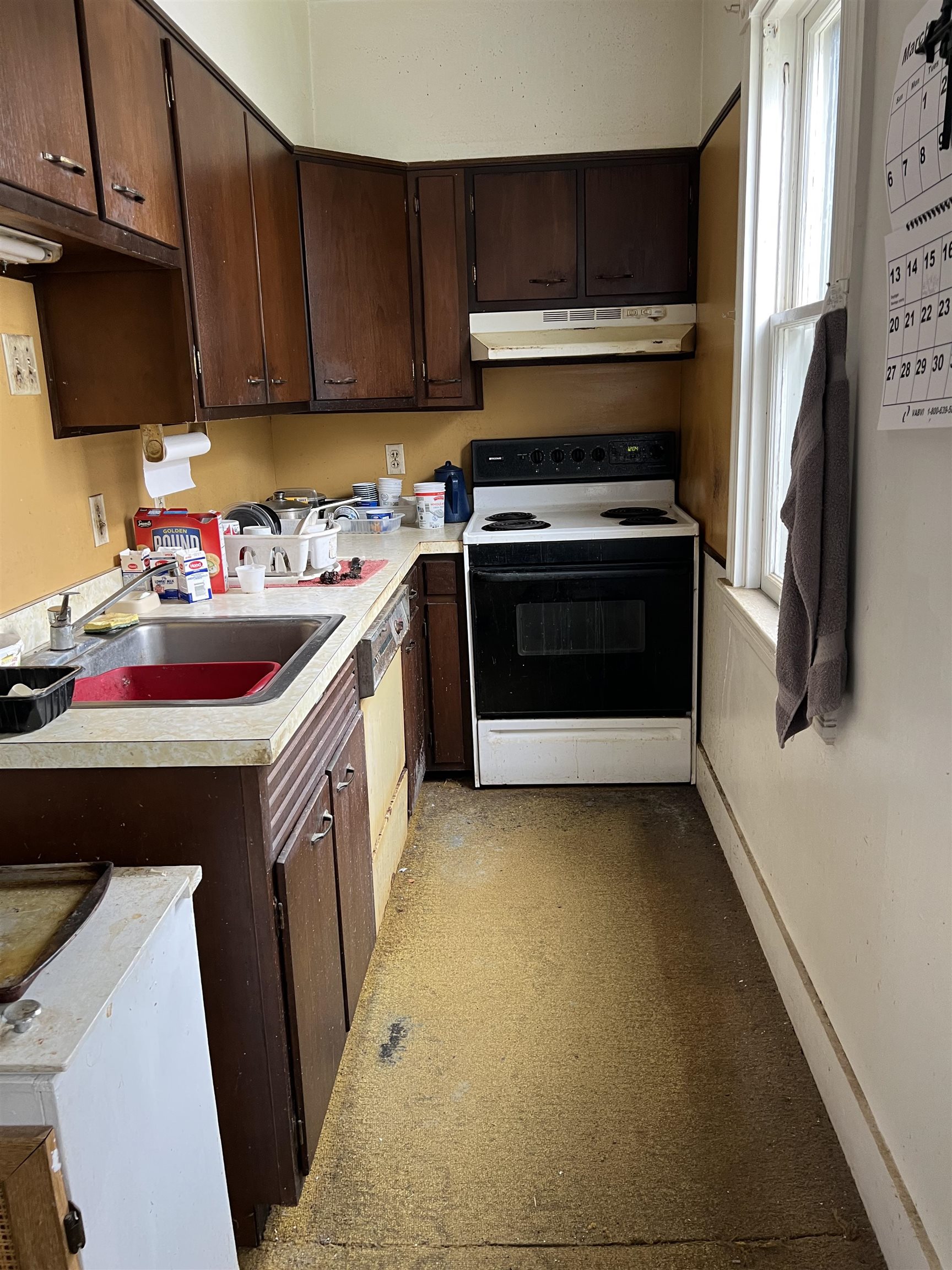 Narrow kitchen with dark wood cabinets, a double sink, and an electric range beside a tall window.