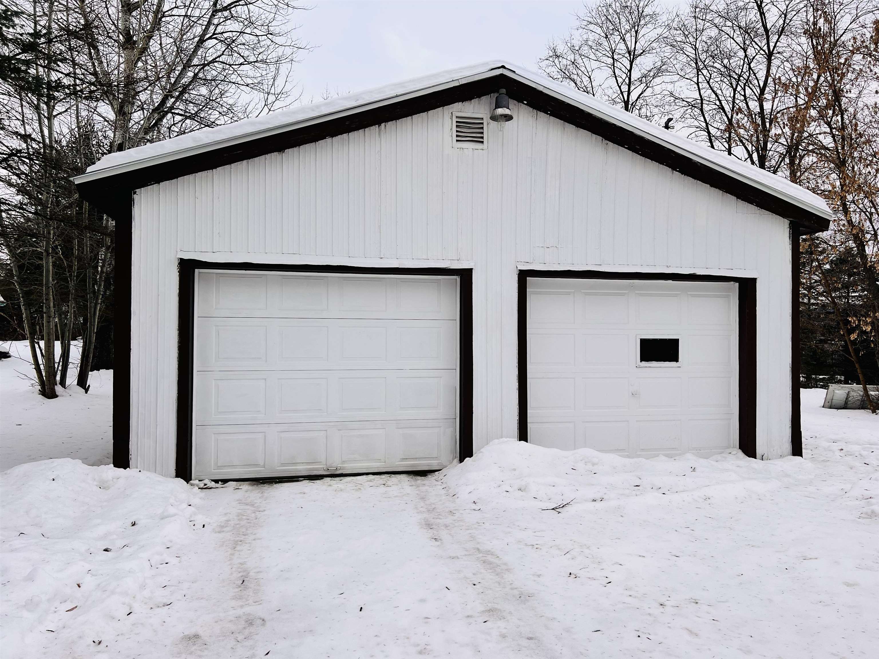 Detached white two-car garage with dark trim, surrounded by snow and leafless trees.