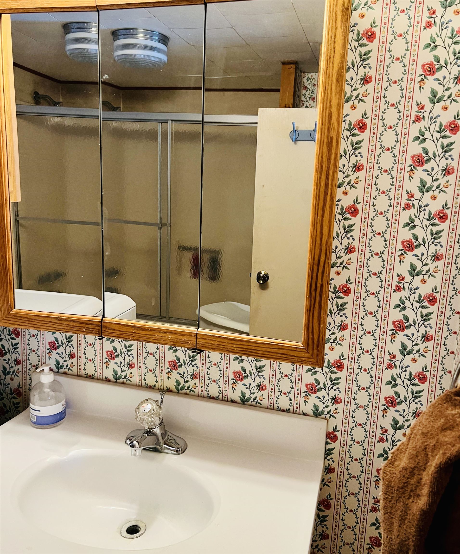 Bathroom vanity with a wood-framed mirror, floral wallpaper, and a tub-shower reflected in the mirror.