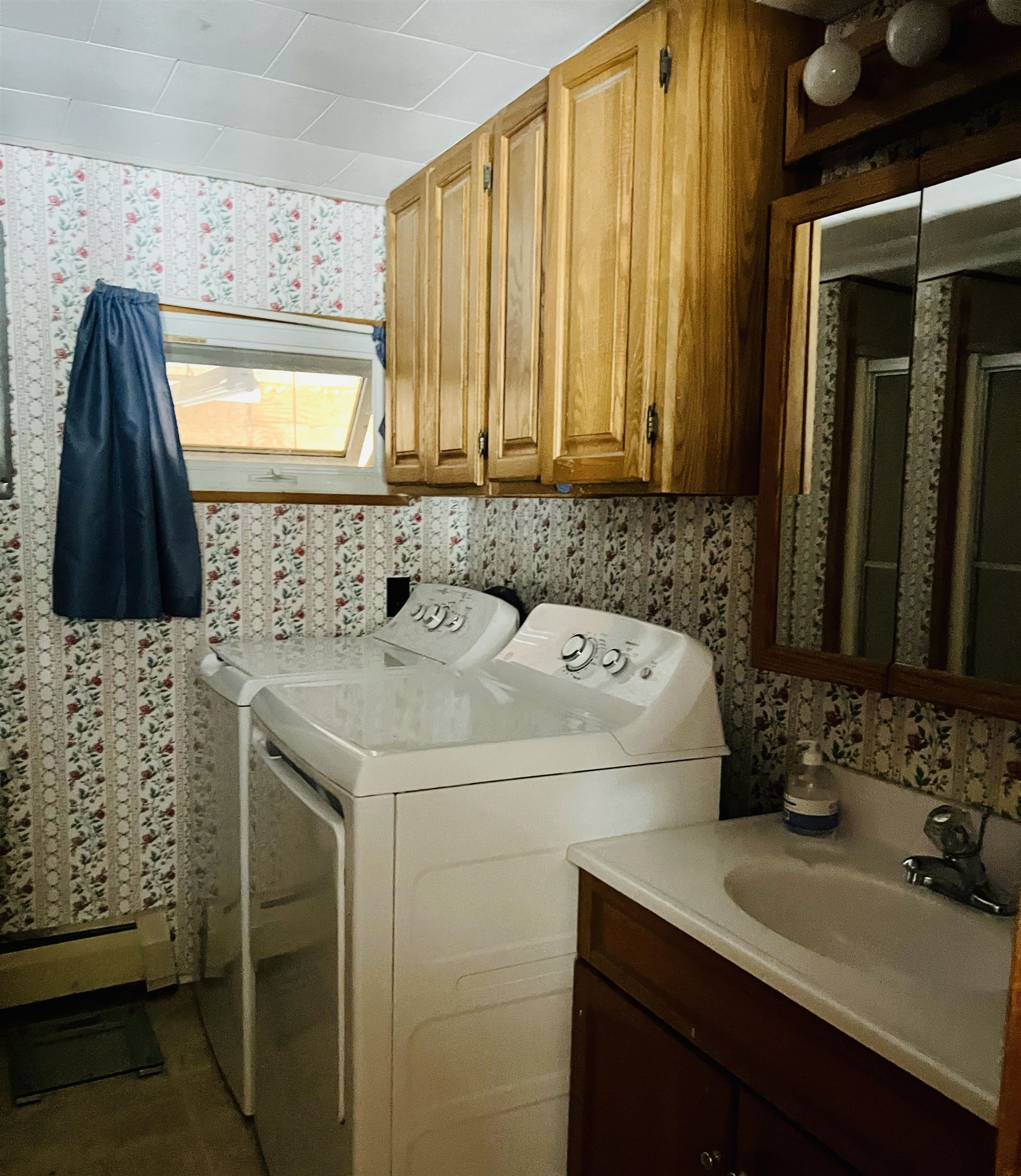 Laundry room with a top-loading washer and dryer, wooden cabinets, floral wallpaper, and a sink.