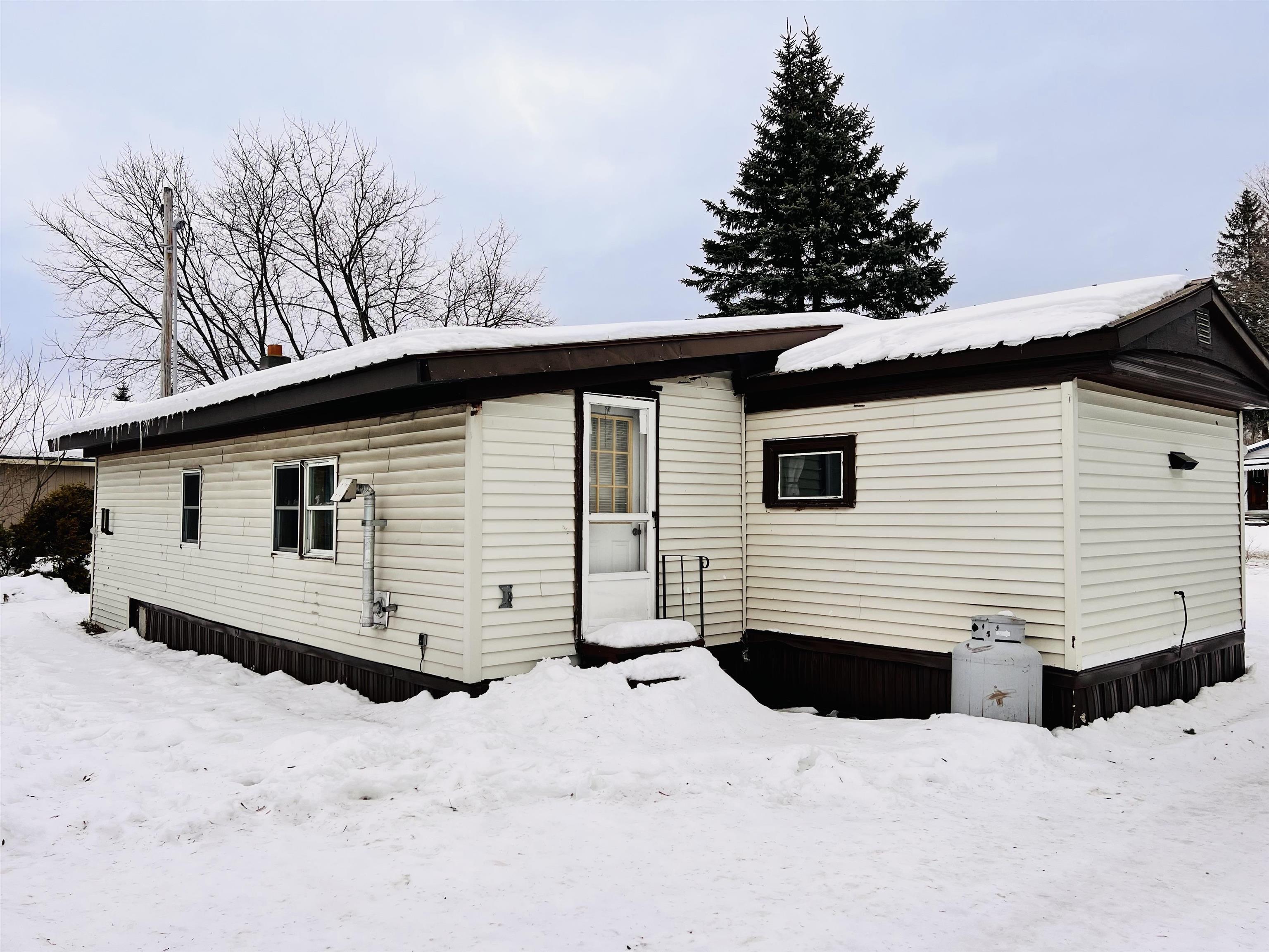 Cream-sided single-story home with dark trim and a snow-covered roof in a snowy yard.