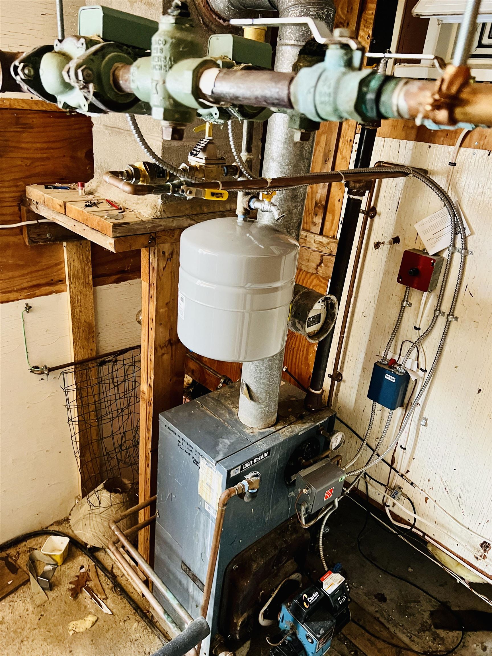 Basement boiler and white expansion tank surrounded by exposed pipes and unfinished wood framing.