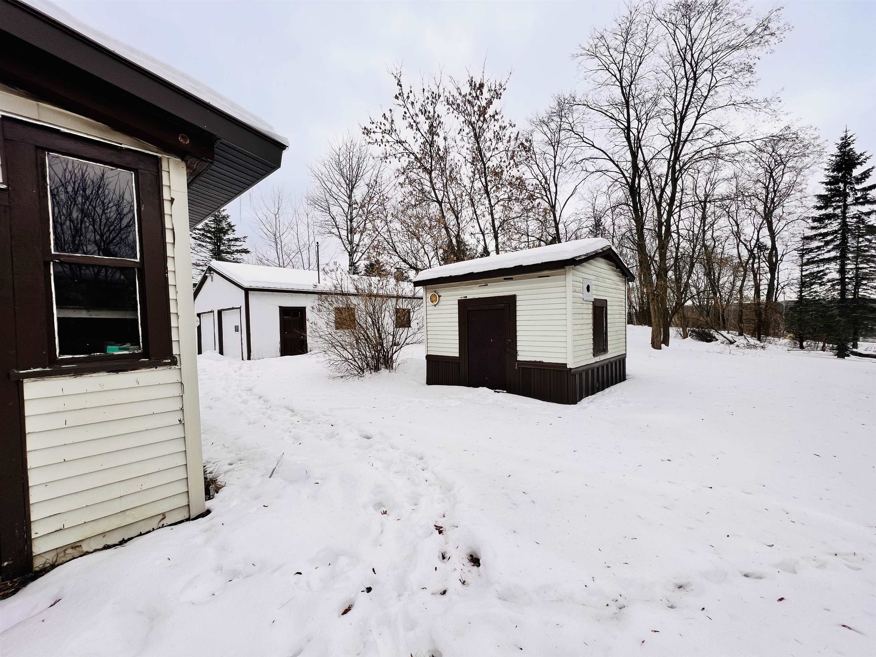 Snowy backyard with a small white outbuilding, detached garage, and bare trees.