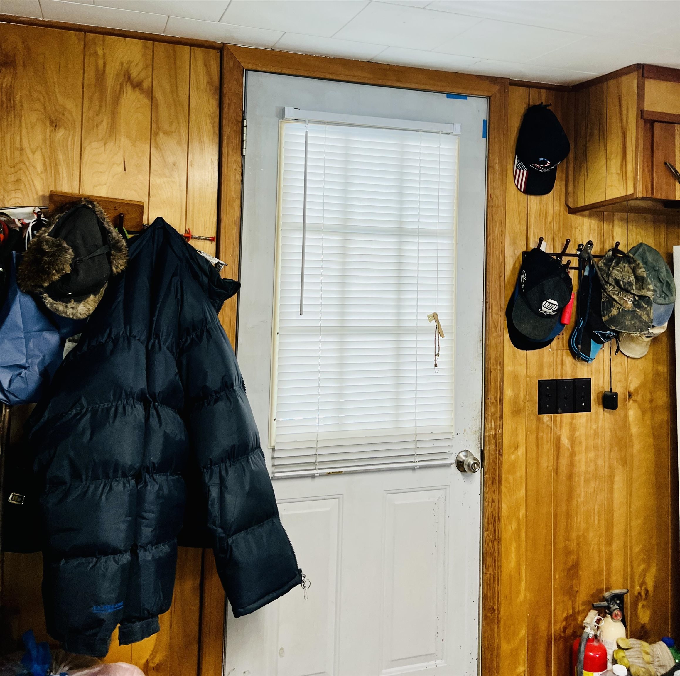 Wood-paneled entryway with a blind-covered door, hanging coats and hats, and a fire extinguisher.