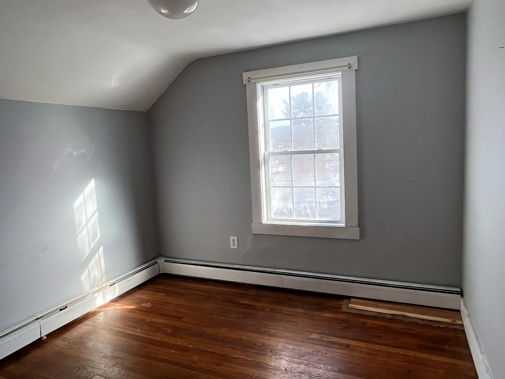 Empty room with a sloped ceiling, hardwood floors, and a sunlit window.