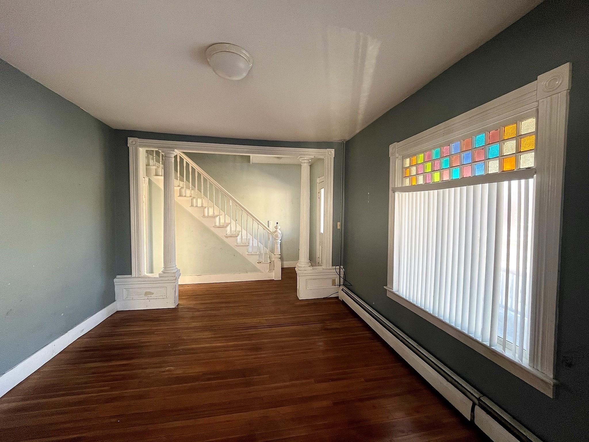 Empty room with hardwood floors, ornate white columns, a staircase, and colorful stained-glass window panes.
