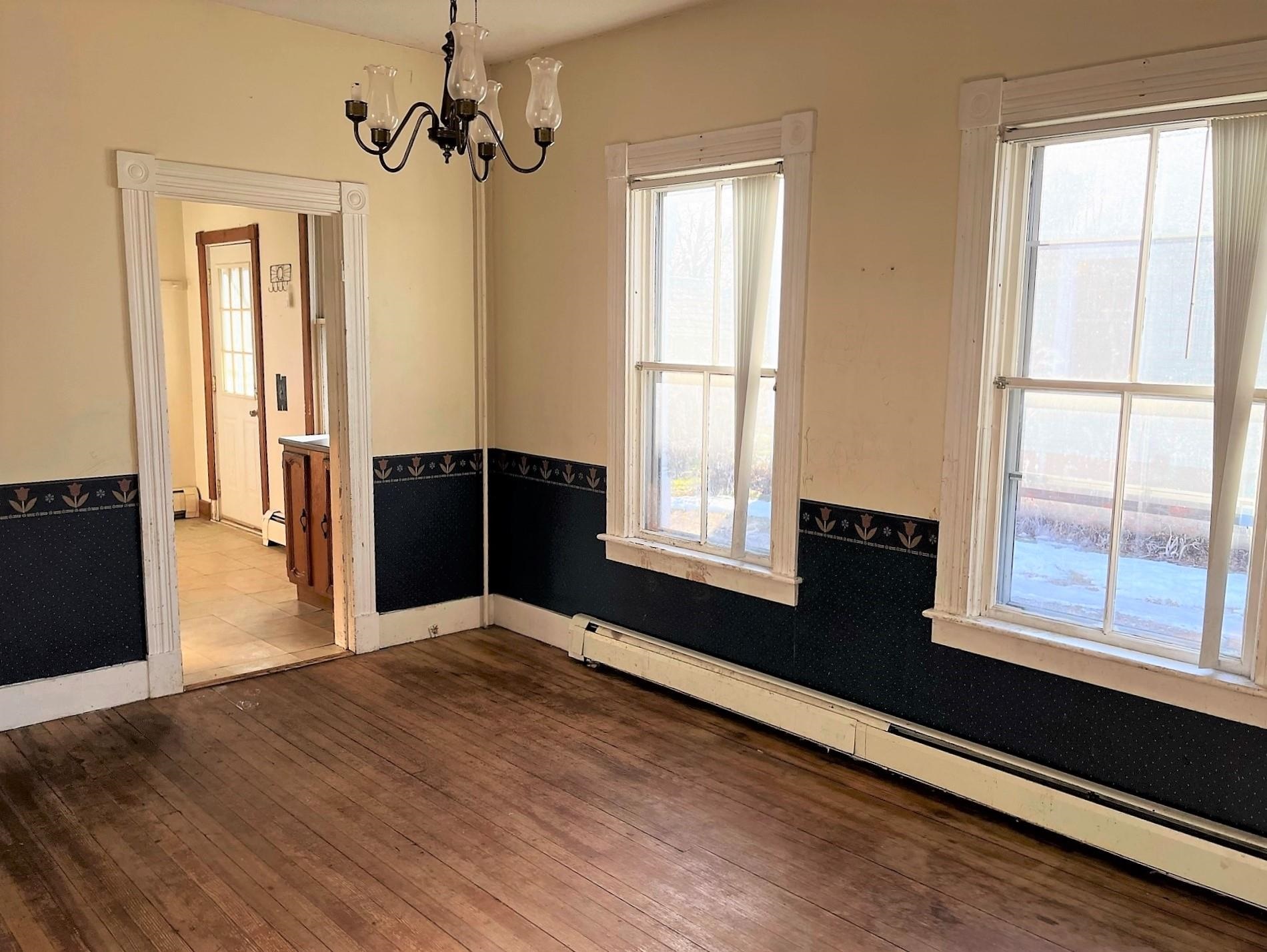 Empty room with hardwood floors, floral wainscoting, two tall windows, and a chandelier.