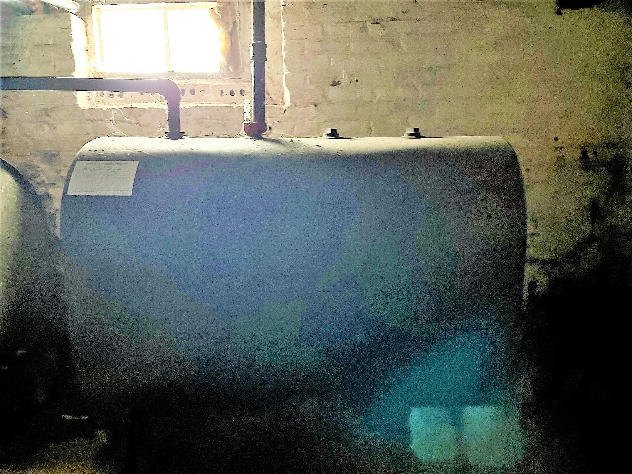 Basement oil tank beneath exposed pipes, with rough masonry walls and a small window.