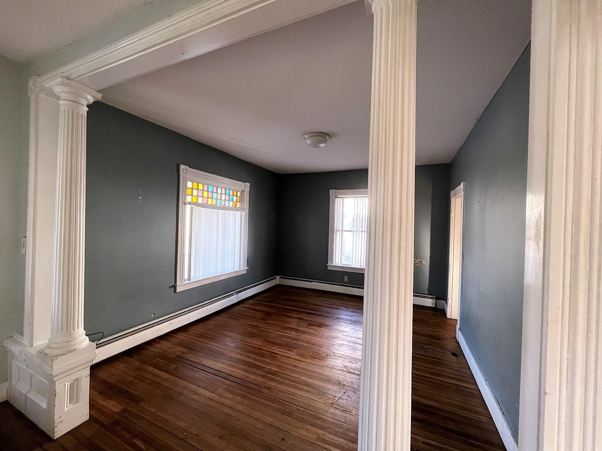 Unfurnished room with dark walls, hardwood floors, colorful transom glass, and white fluted columns.
