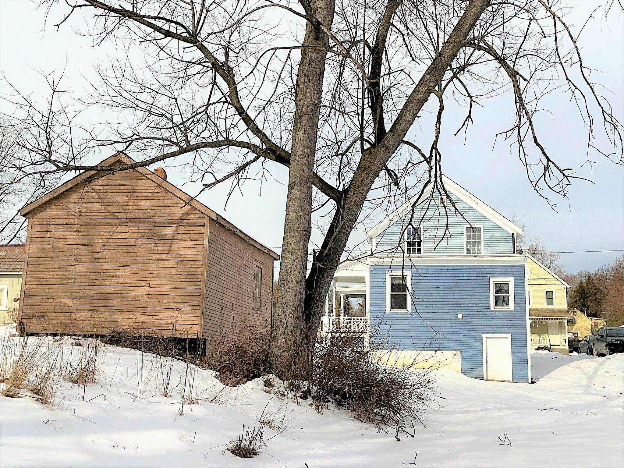 Blue-sided house and wooden outbuilding stand behind a large leafless tree in a snowy yard.