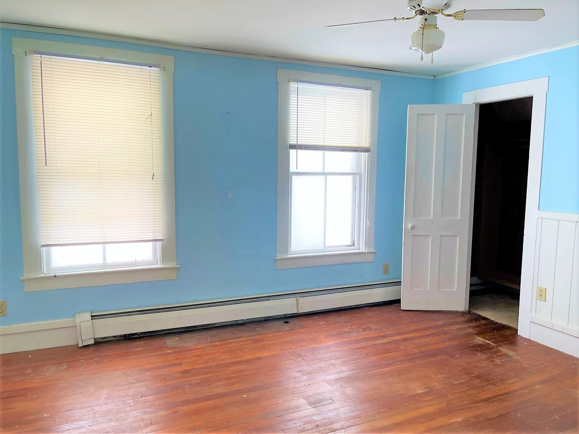 Empty blue room with two blinded windows, a ceiling fan, and worn hardwood floors.