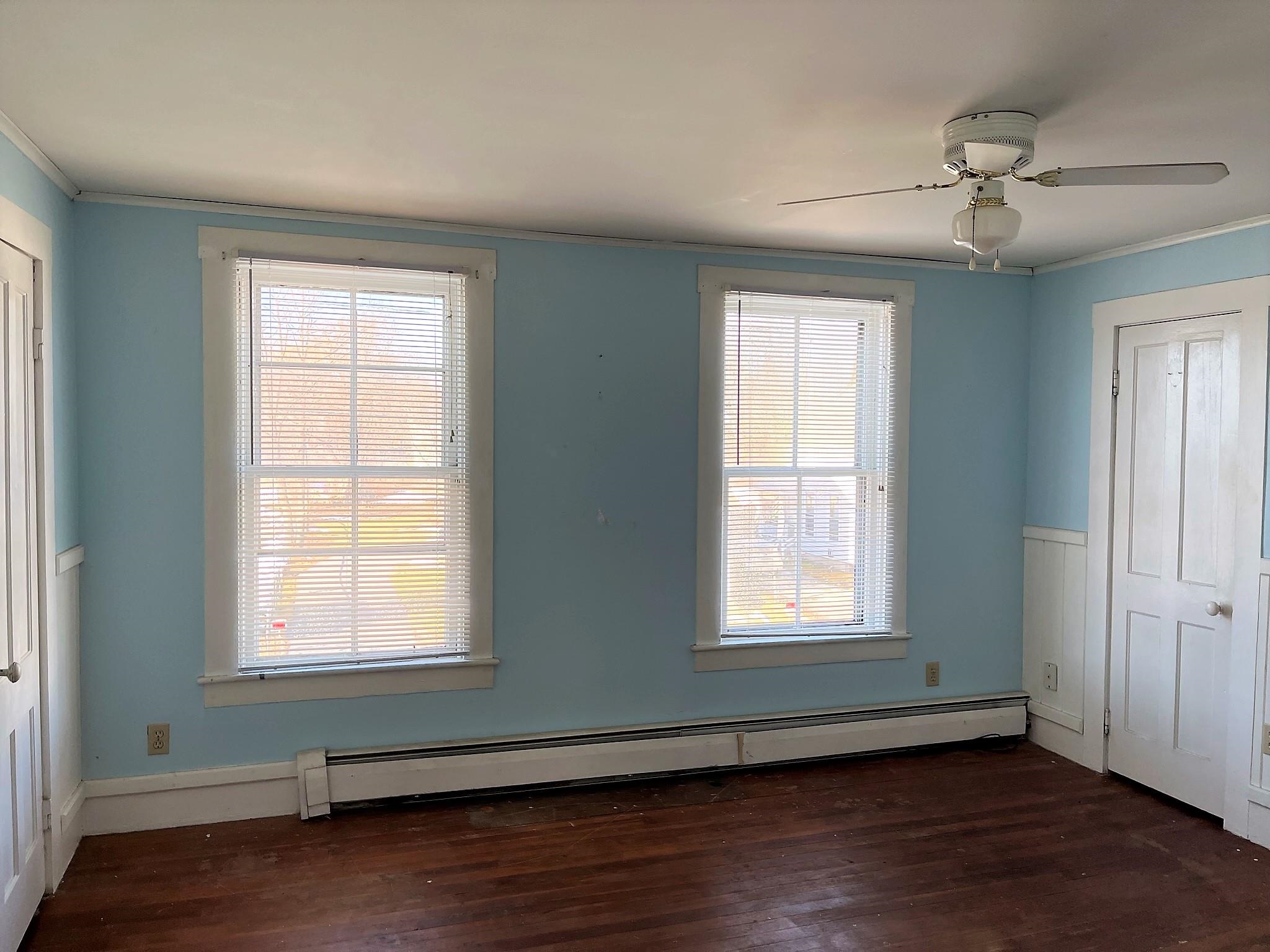 Empty blue room with dark wood floors, two blinded windows, ceiling fan, and baseboard radiator.