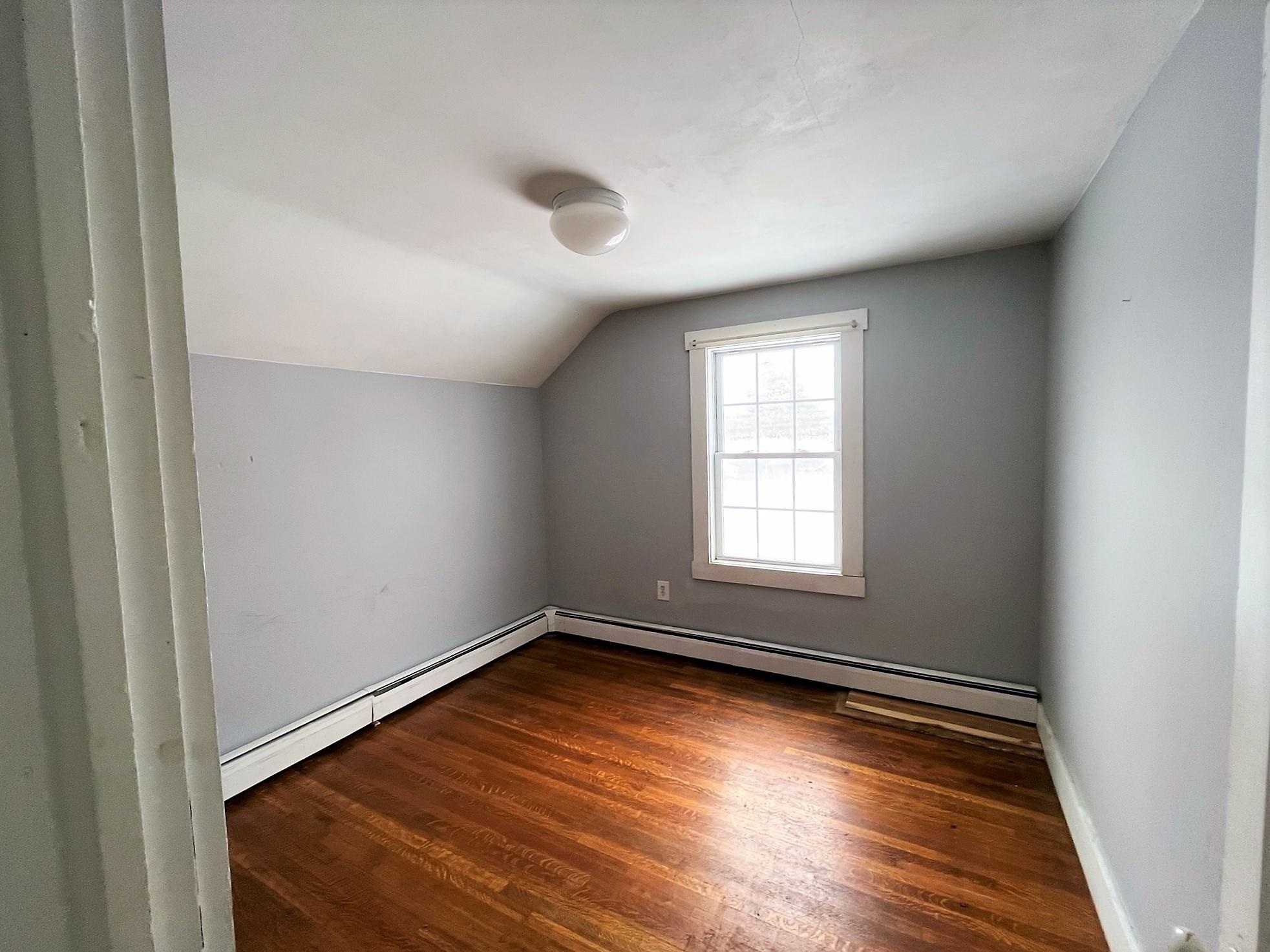 Empty room with hardwood floors, gray walls, and a sloped ceiling above a single window.