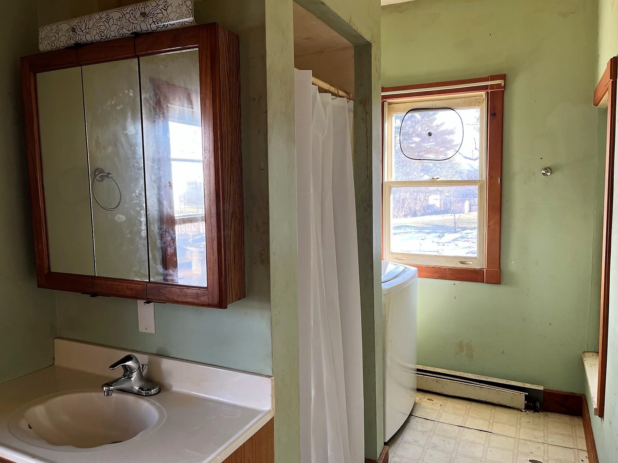 Compact bathroom with a wooden mirrored vanity, shower curtain, and washing machine beside a window.