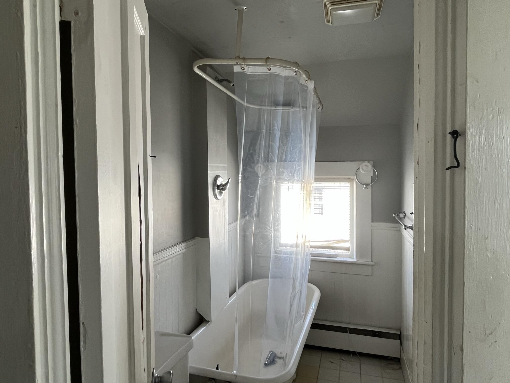 Bathroom with a white bathtub, clear shower curtain, tiled floor, and window above the tub.
