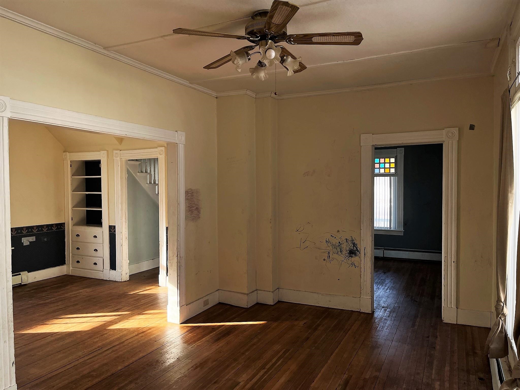 Empty room with worn hardwood floors, ornate doorways, built-in shelving, and a ceiling fan.