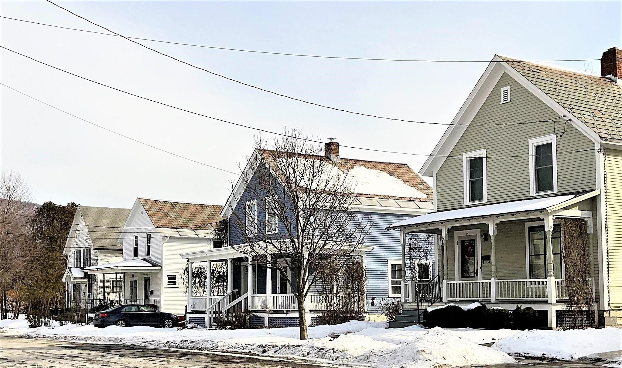 Sage-green two-story house with a covered porch, snow-covered yard, and neighboring homes.