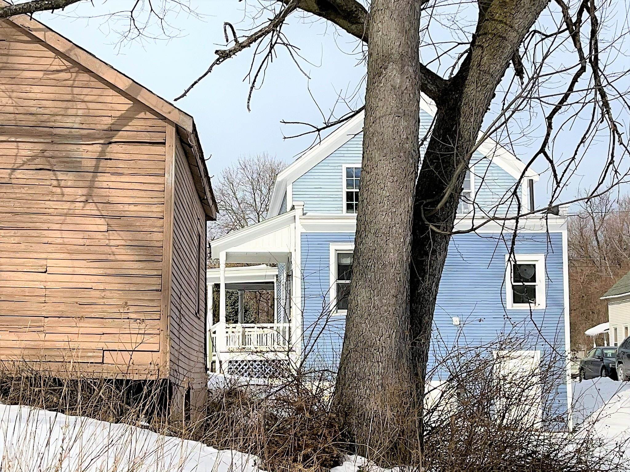 Blue house with a covered porch, partly hidden behind a large leafless tree in snow.