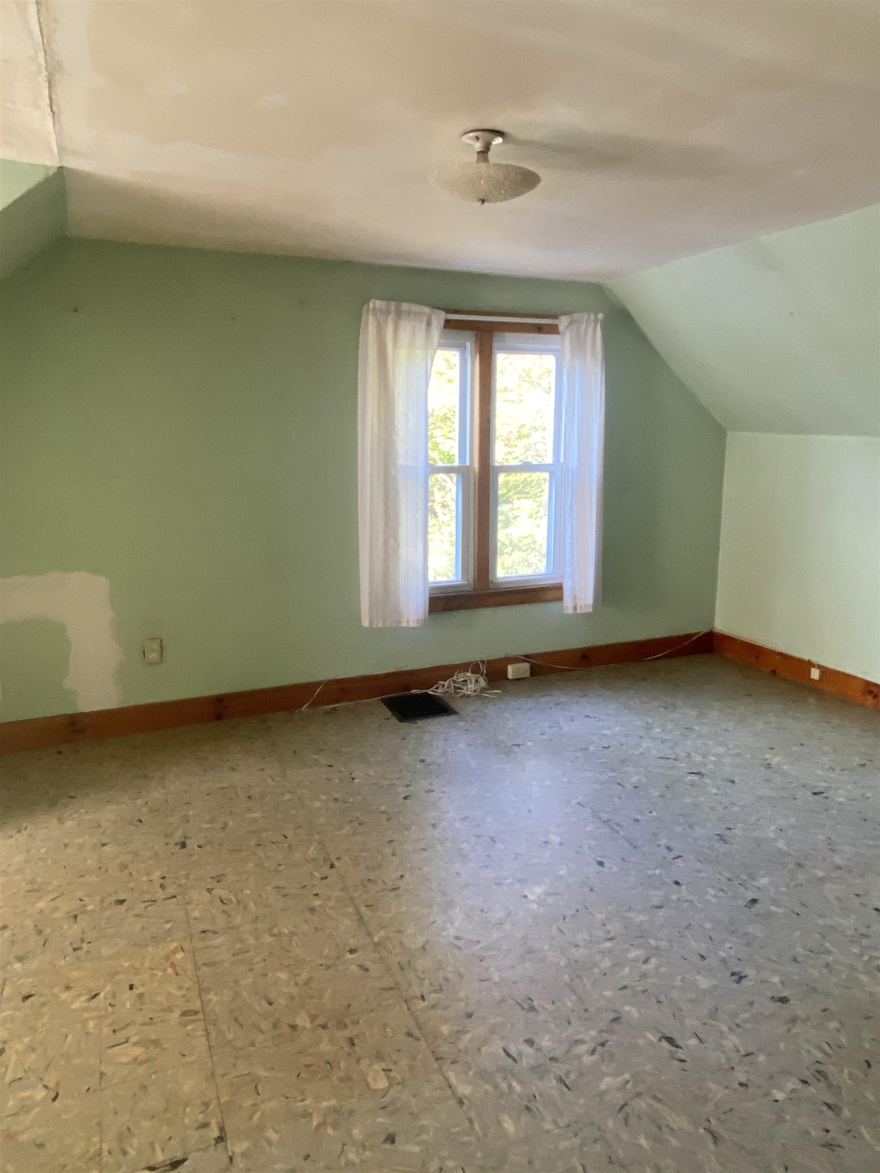 Empty attic room with green walls, sloped ceiling, unfinished floor, and a curtained window.