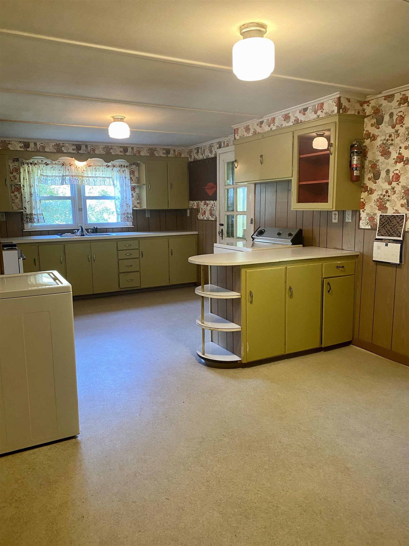 Vintage kitchen with olive-green cabinets, a curved peninsula, wood paneling, and floral wallpaper.
