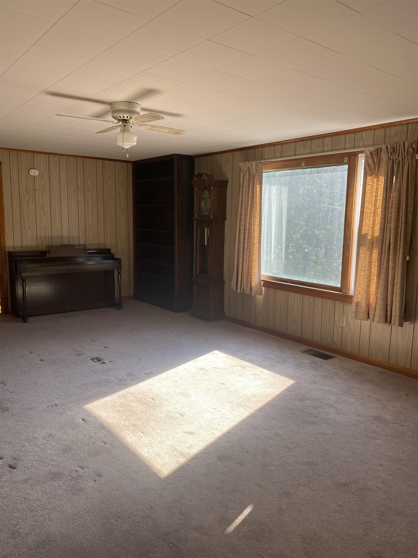 Carpeted room with wood paneling, a piano, grandfather clock, and sunlight streaming through a large window.