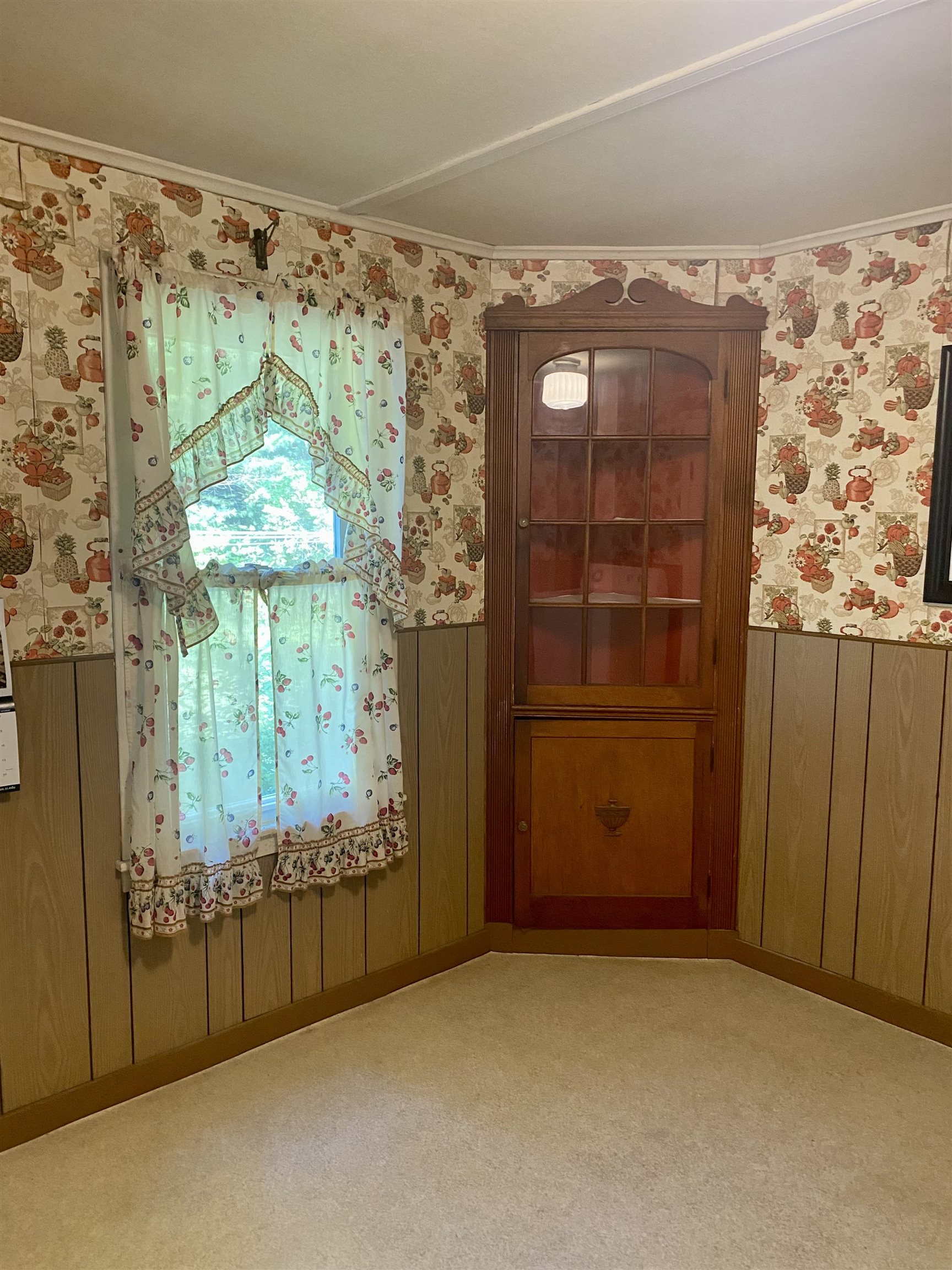 Interior corner with a tall wooden glass-front cabinet beside a window framed by fruit-patterned curtains.