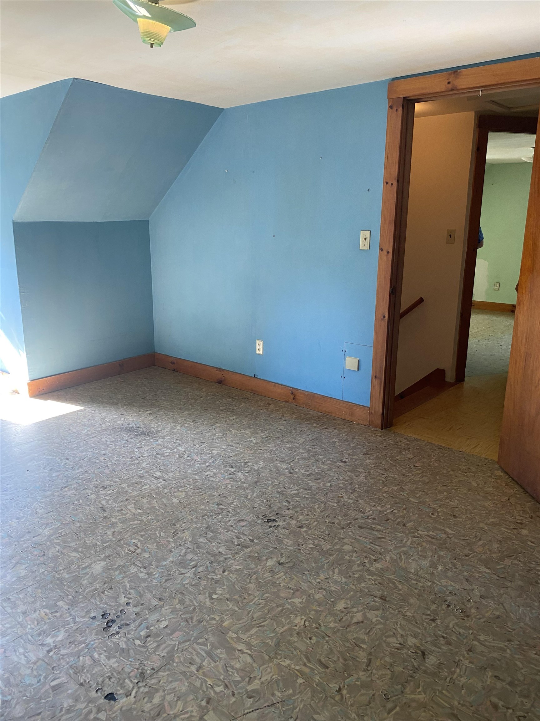 Empty blue-walled room with a sloped ceiling, exposed subfloor, and wood-trimmed doorway.