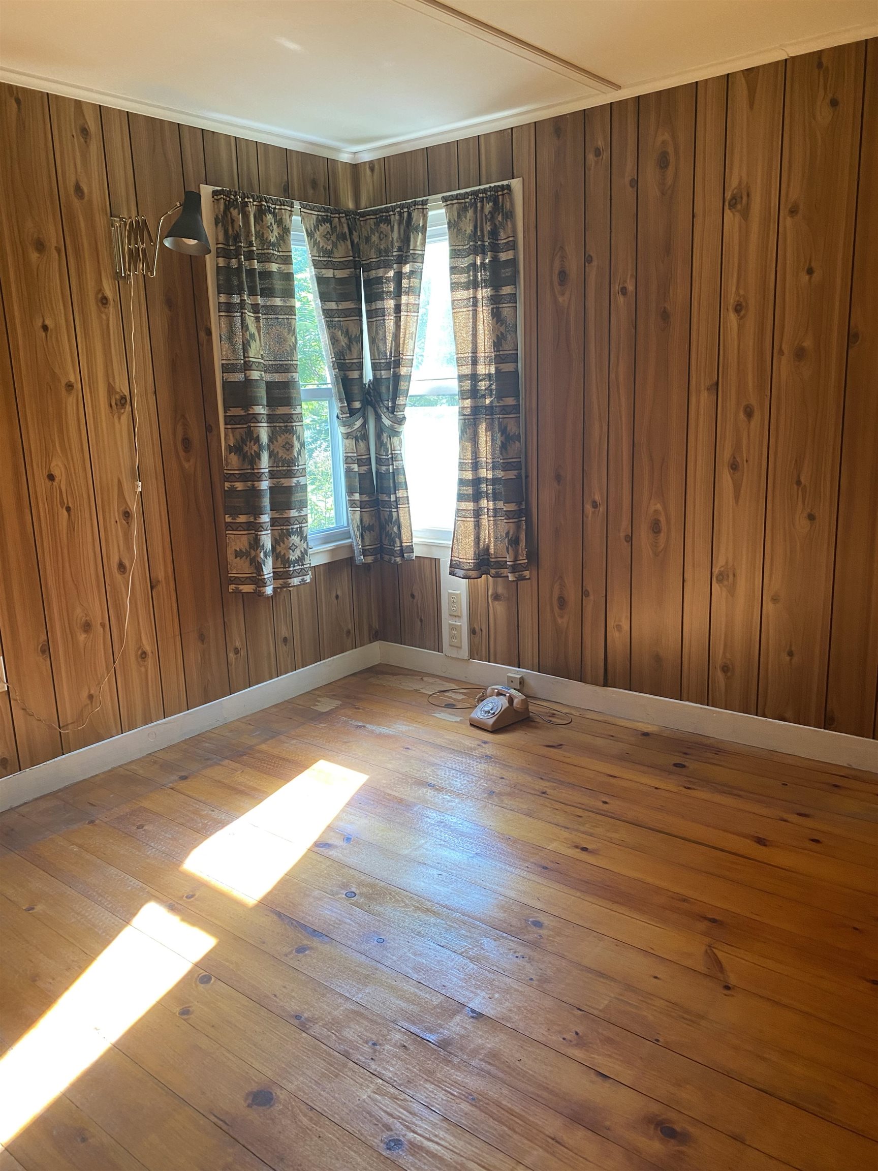 Mostly empty wood-paneled room with plank floors, a curtained corner window, and sunlight across the floor.