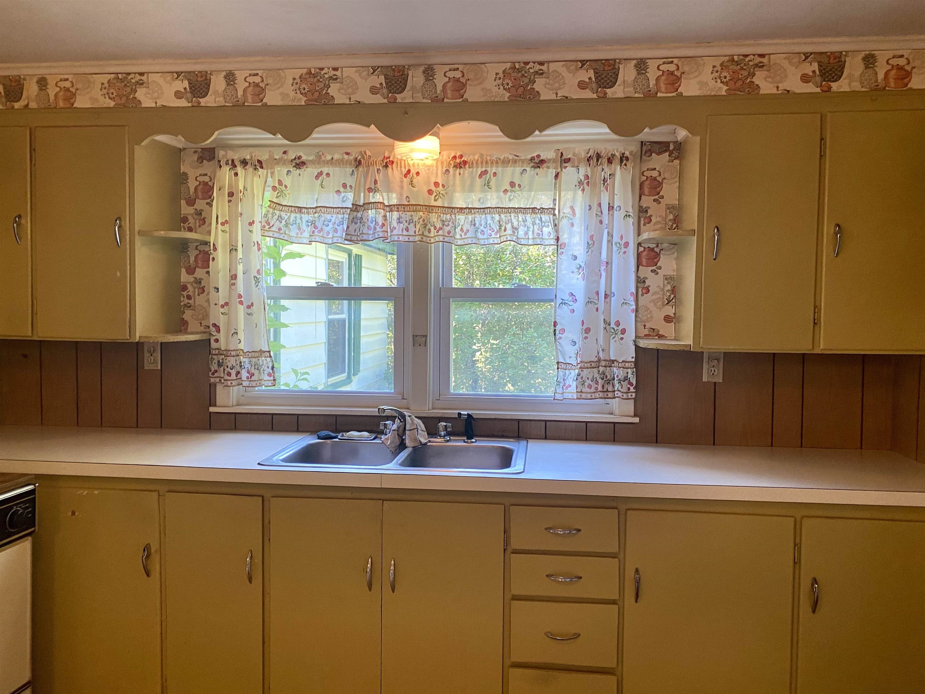 Kitchen with a double-basin sink beneath a window framed by floral curtains and mustard-yellow cabinets.