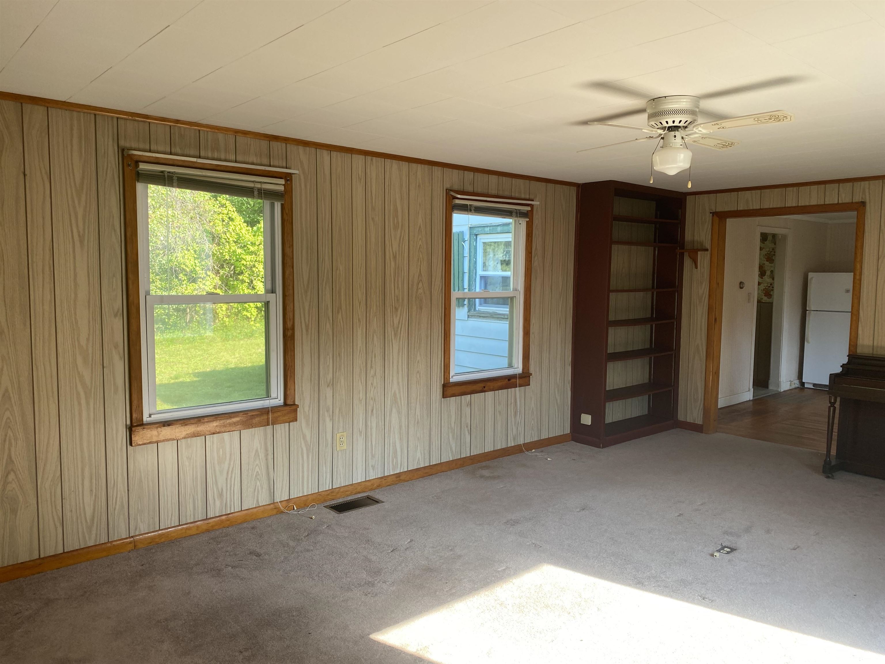 Empty carpeted room with wood paneling, built-in shelves, and a doorway to the kitchen.