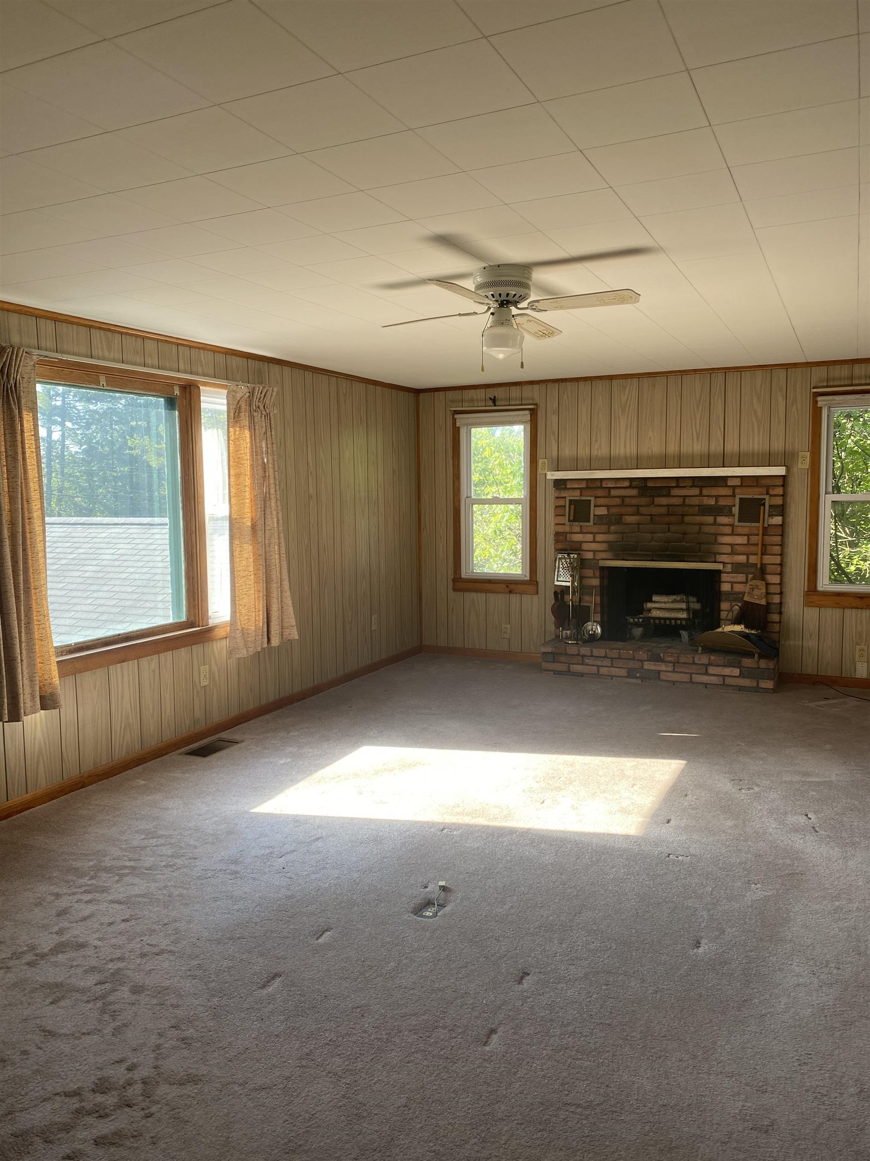 Unfurnished living room with a brick fireplace, wood-paneled walls, and large windows.