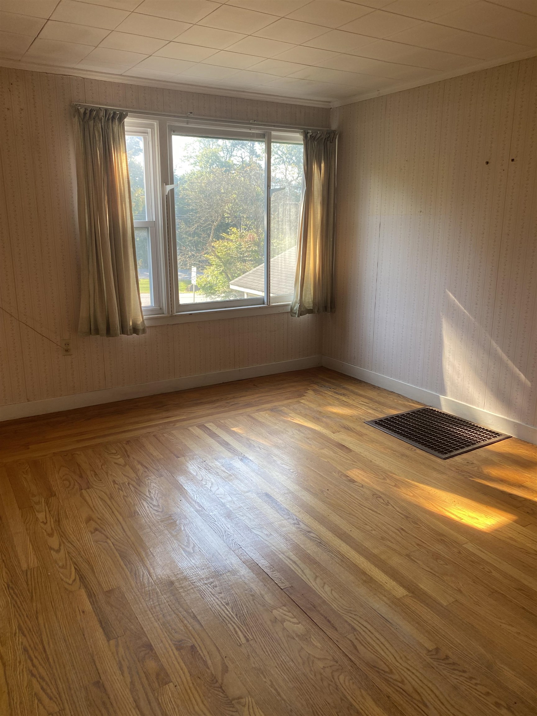 Empty room with hardwood floors, patterned wallpaper, and sunlight streaming through a curtained window.