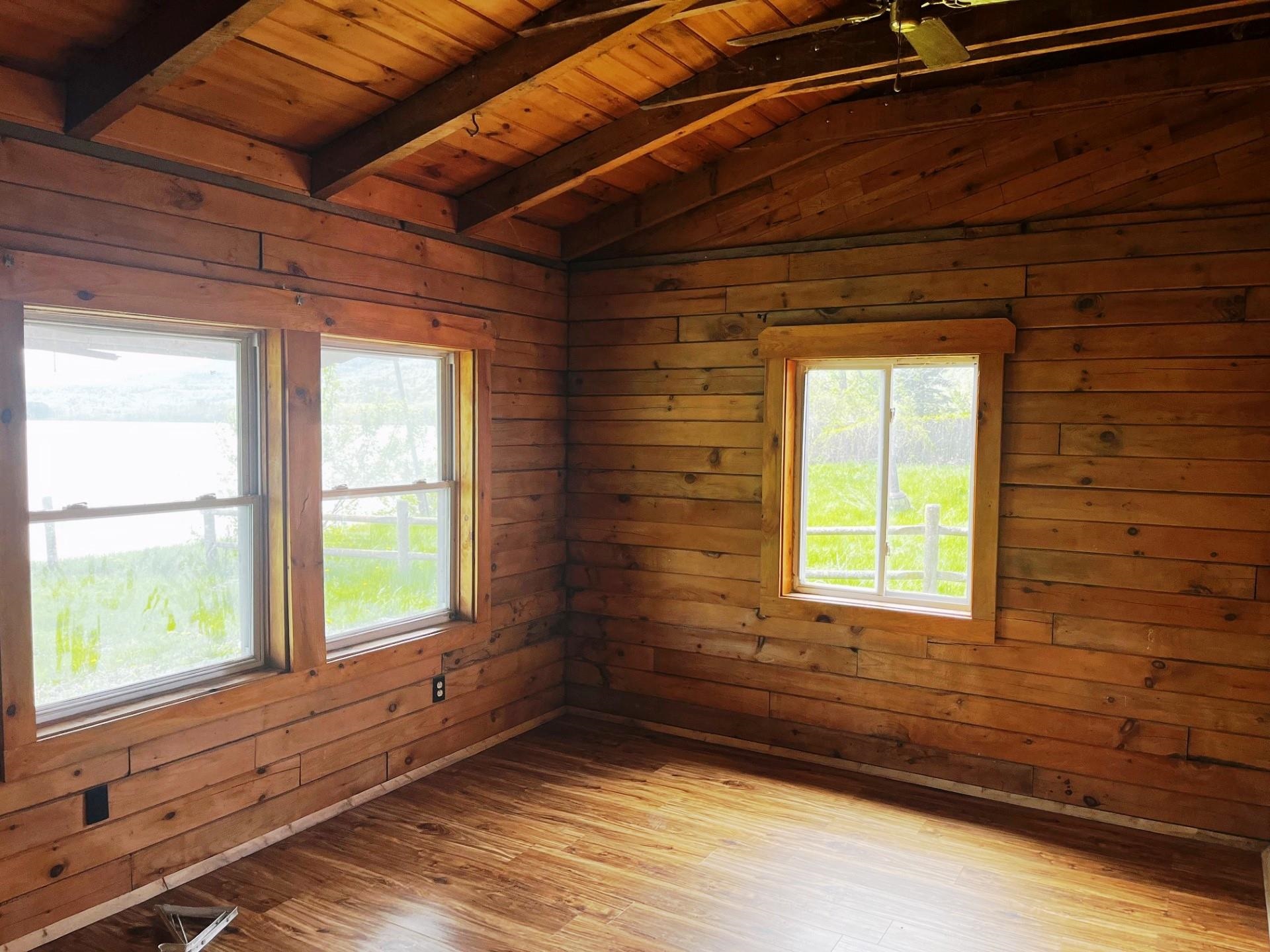 Empty wood-paneled room with a vaulted beam ceiling, three windows, and wood-look flooring.