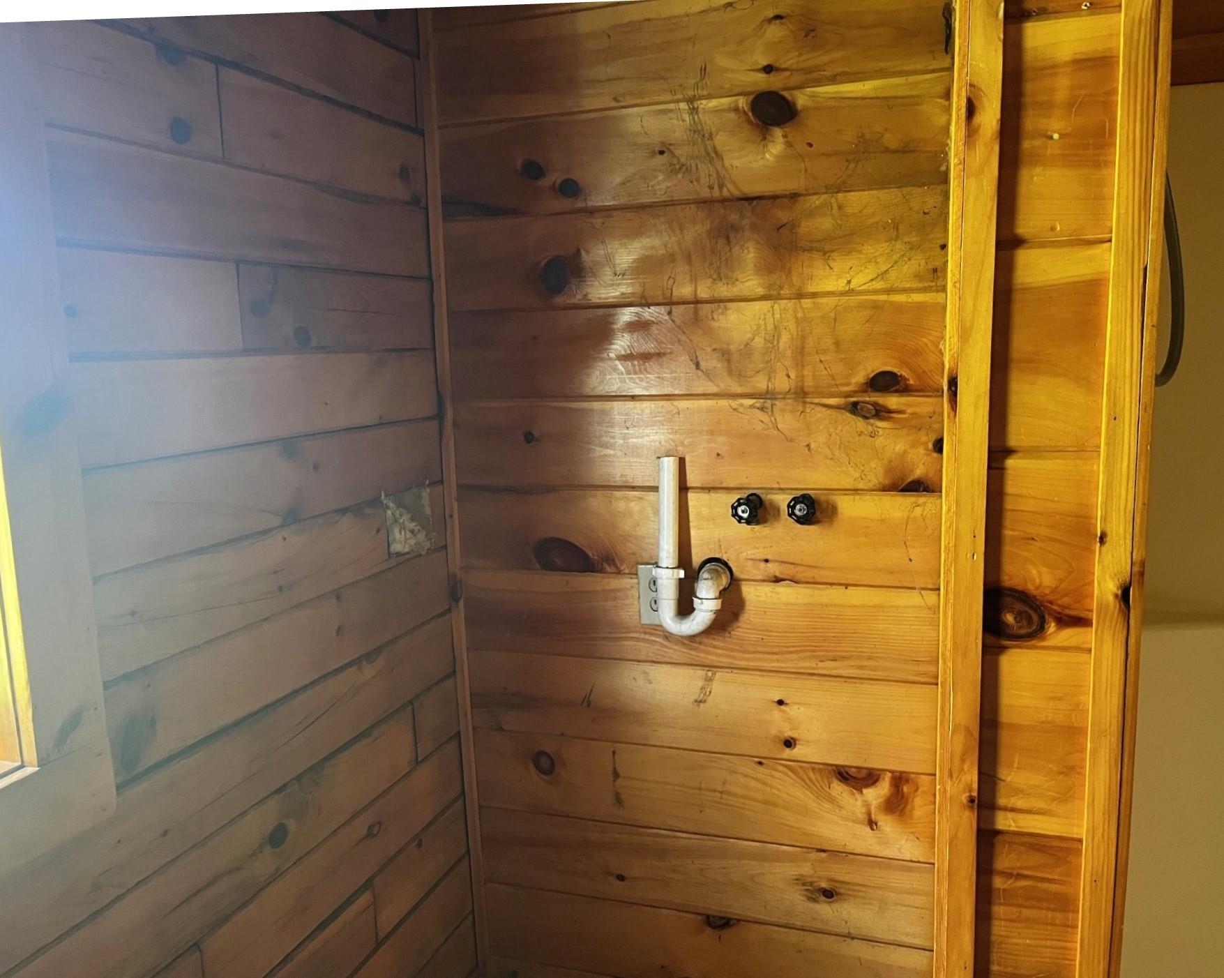 Wood-paneled interior with exposed plumbing, two wall controls, and a white drainpipe.