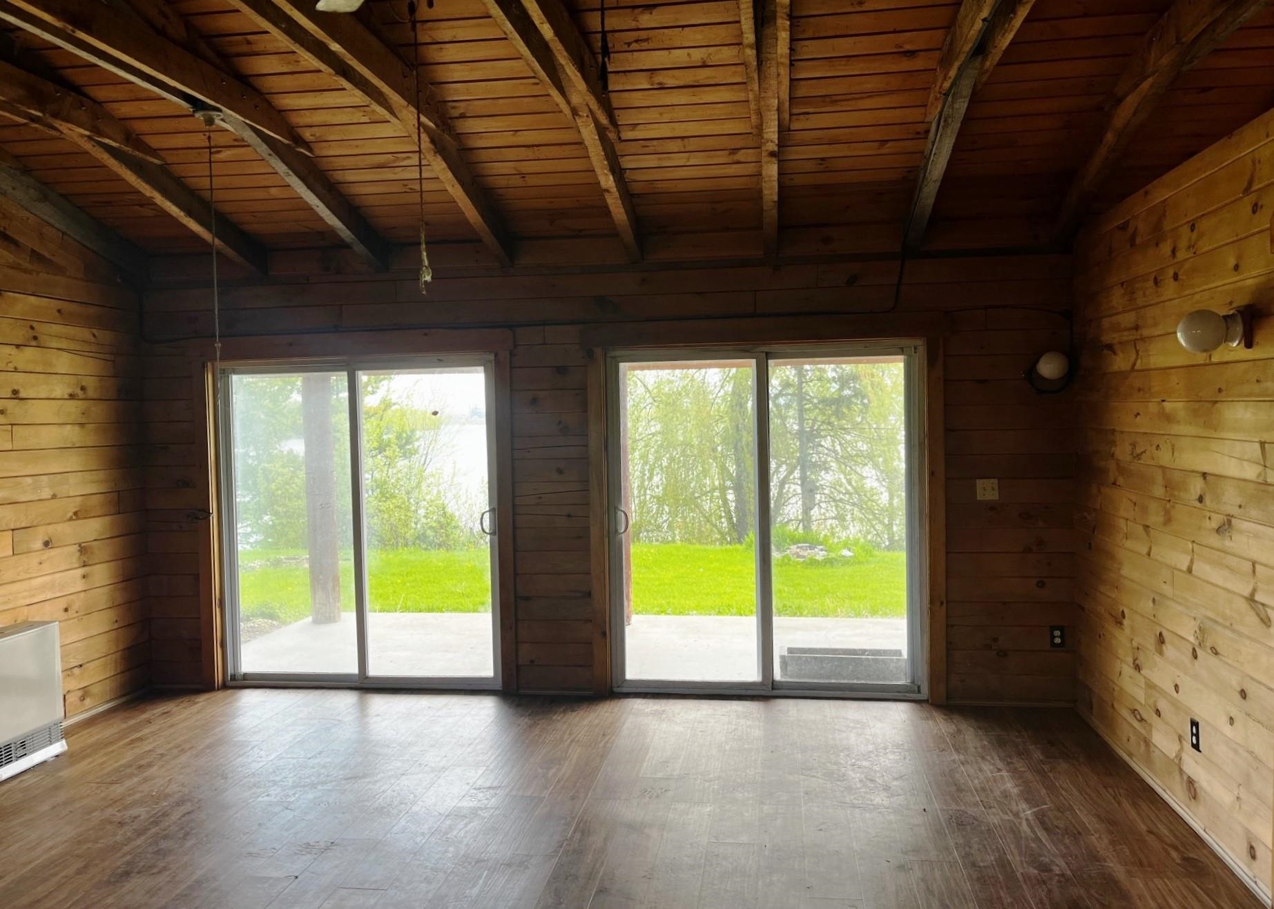 Empty wood-paneled room with exposed beams and sliding glass doors overlooking a lawn and water.