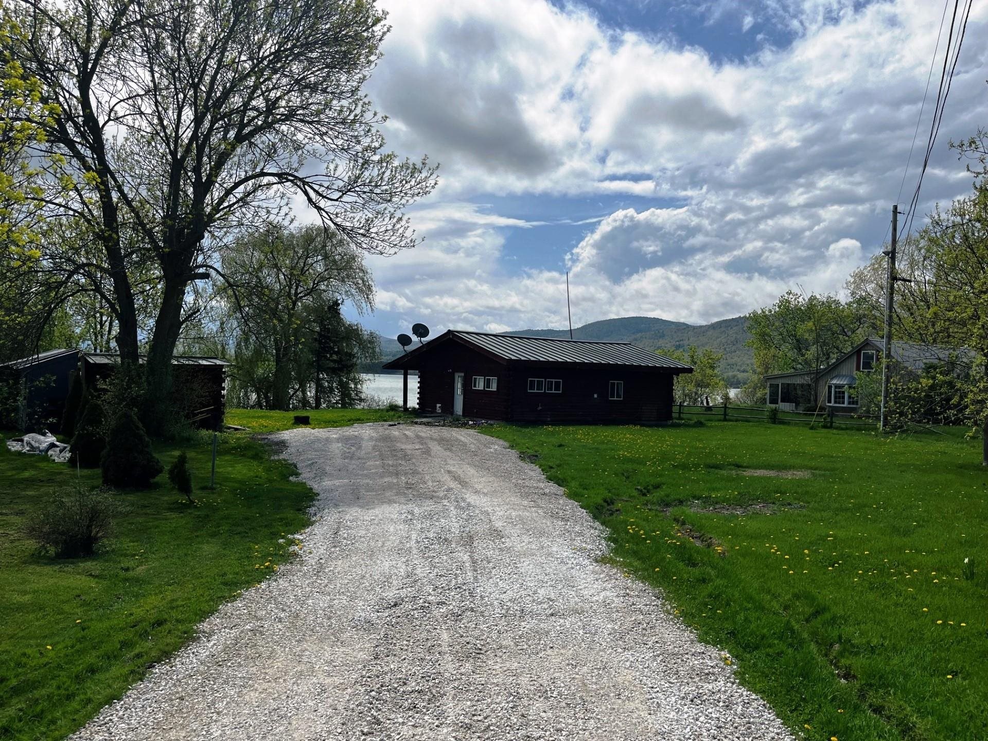 Dark single-story house at the end of a gravel driveway, with lake and wooded hills behind it.