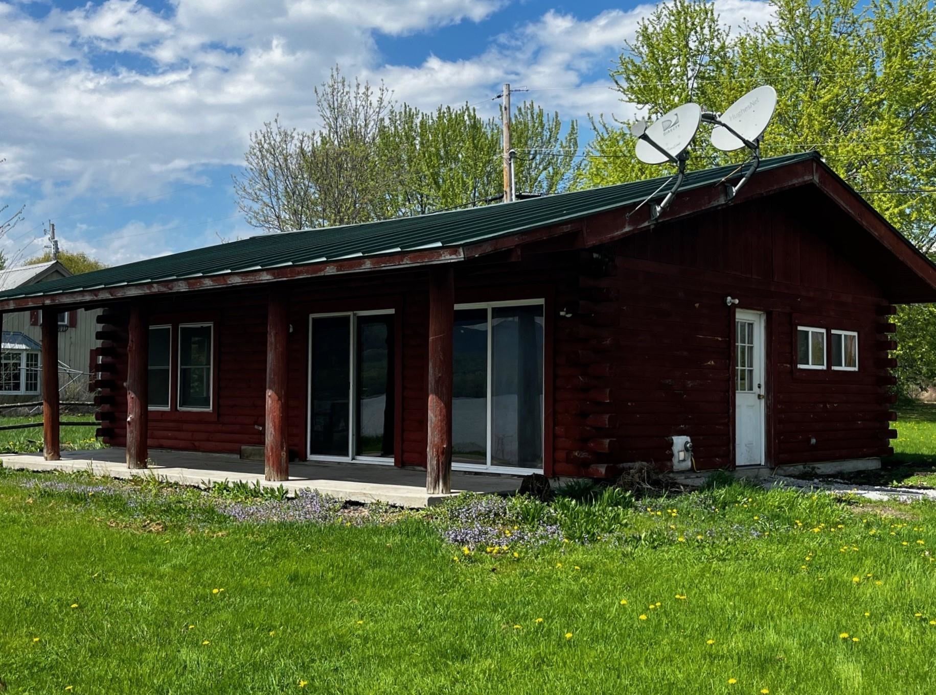 Red log cabin with a green metal roof, covered porch, and satellite dishes above a flower-dotted lawn.