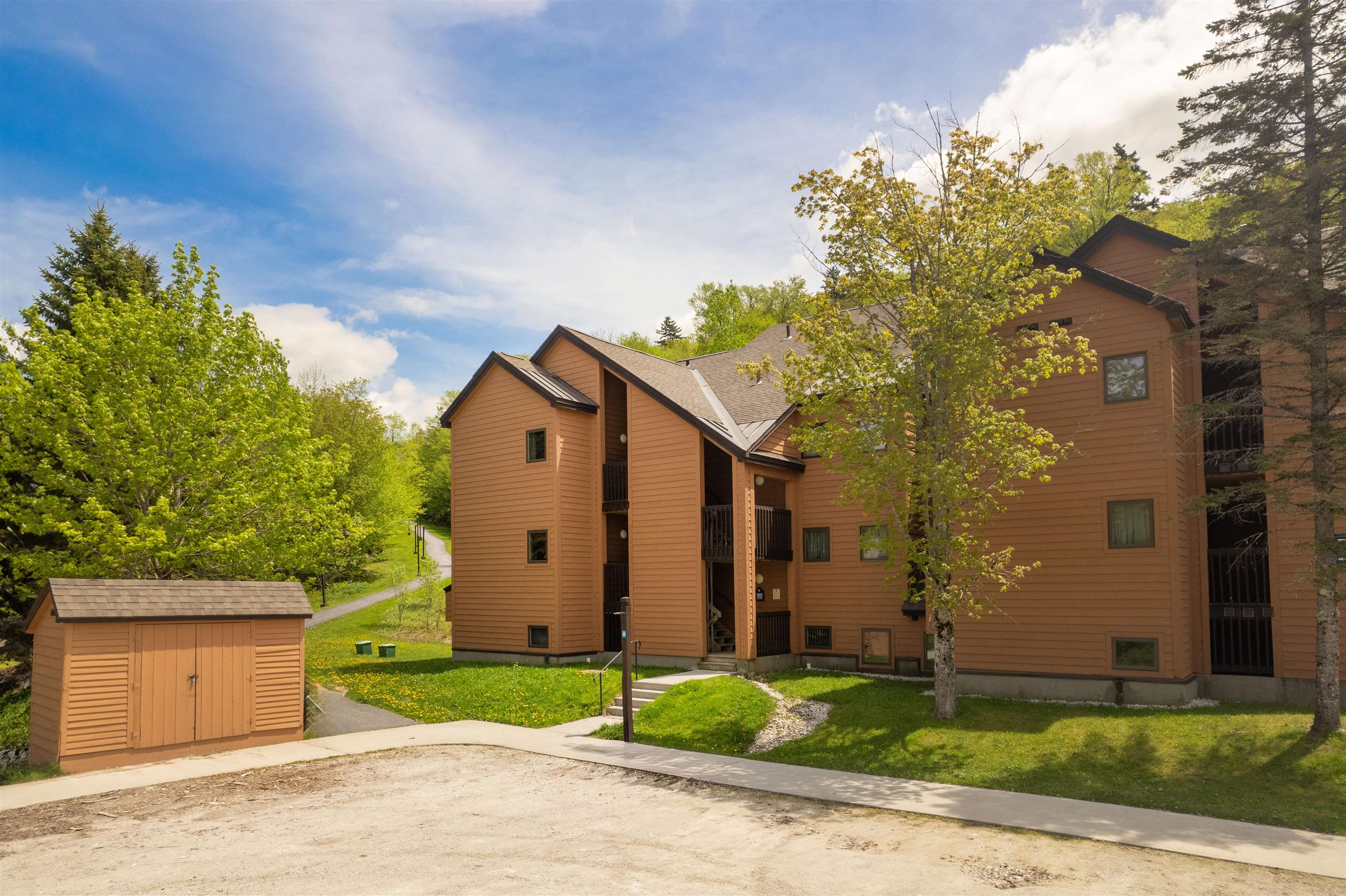 Brown condominium building with gabled roofs and balconies among leafy trees.