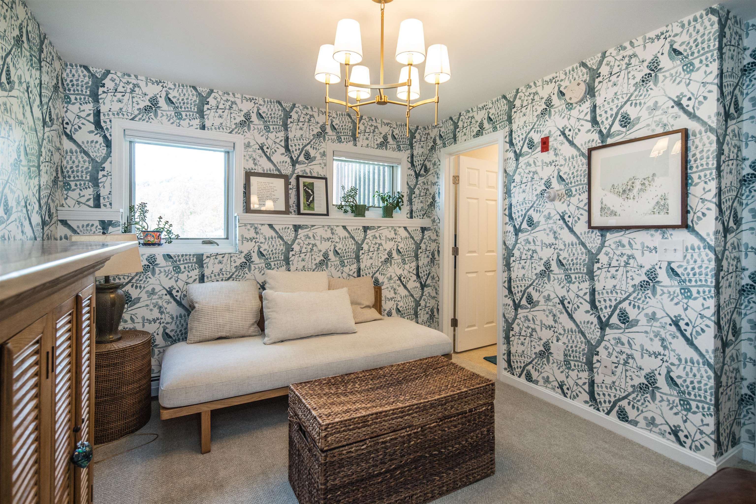 Sitting room with botanical wallpaper, a cushioned daybed, woven trunk, and brass chandelier.