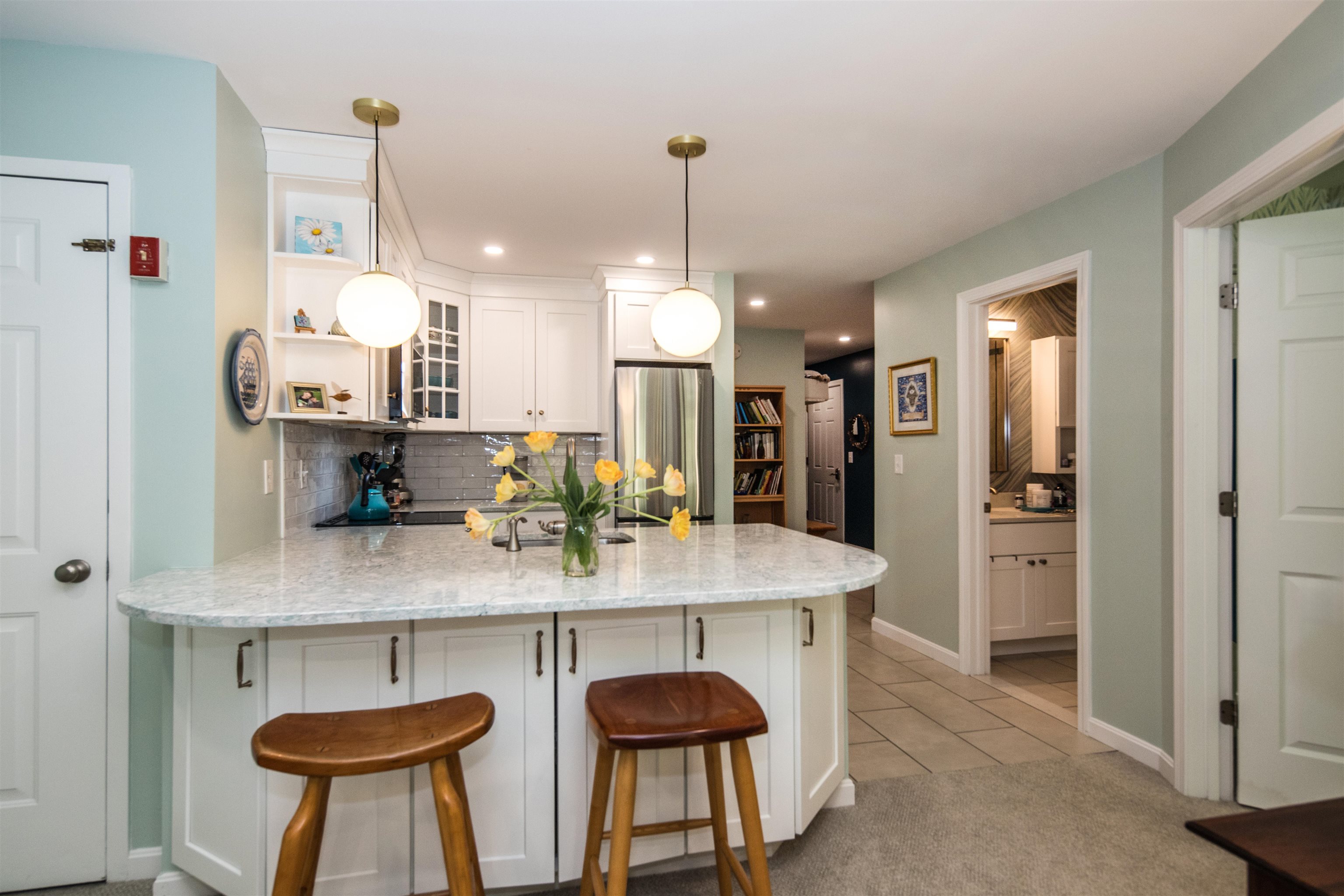 Furnished kitchen with a stone-topped island, wooden stools, and yellow flowers beneath globe pendant lights.