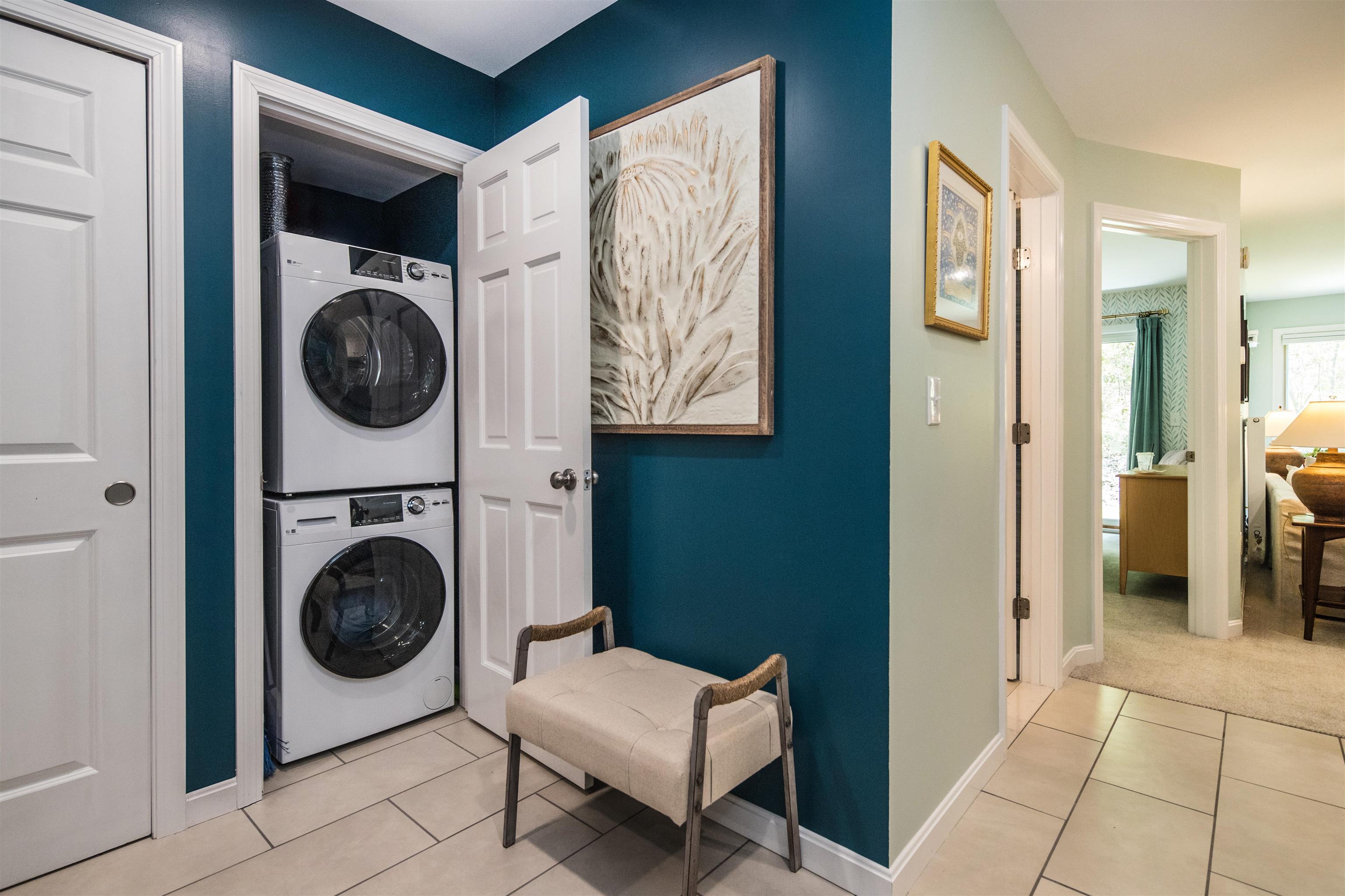 Laundry area with stacked washer and dryer, teal accent wall, and upholstered bench.