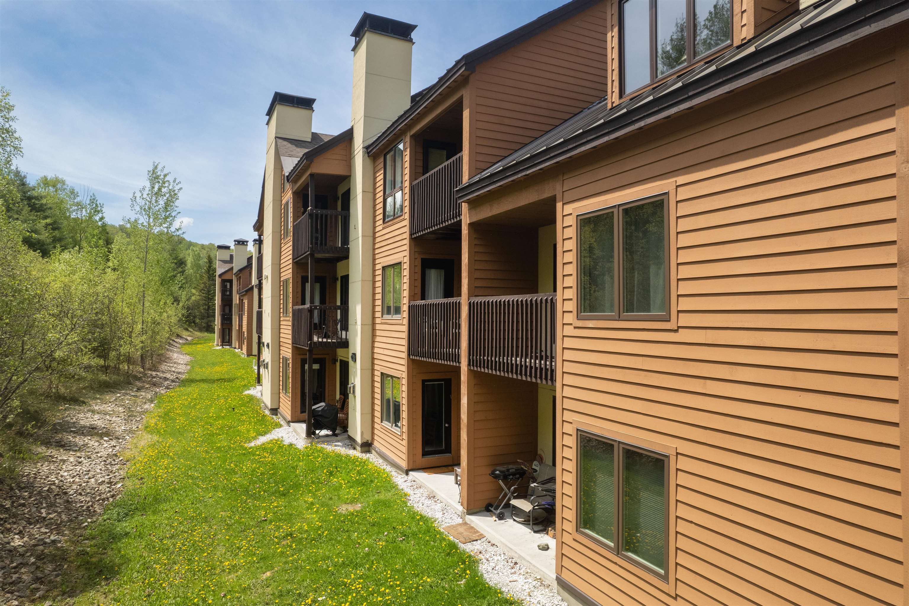 Tan condominium building with balconies beside a flower-dotted lawn and wooded grounds.