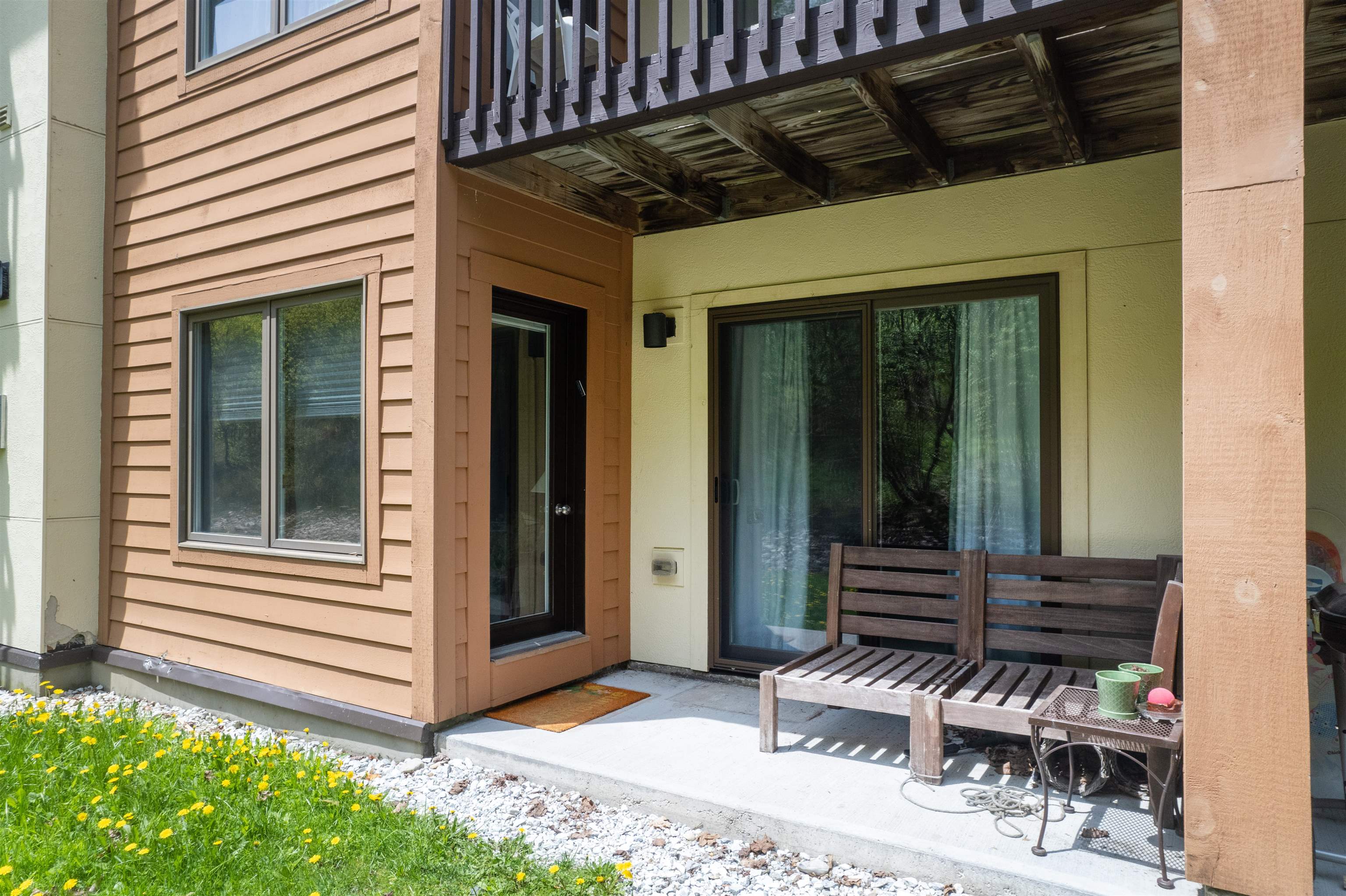 Condominium patio beneath an upper balcony, with a wooden bench, small table, and sliding glass doors.