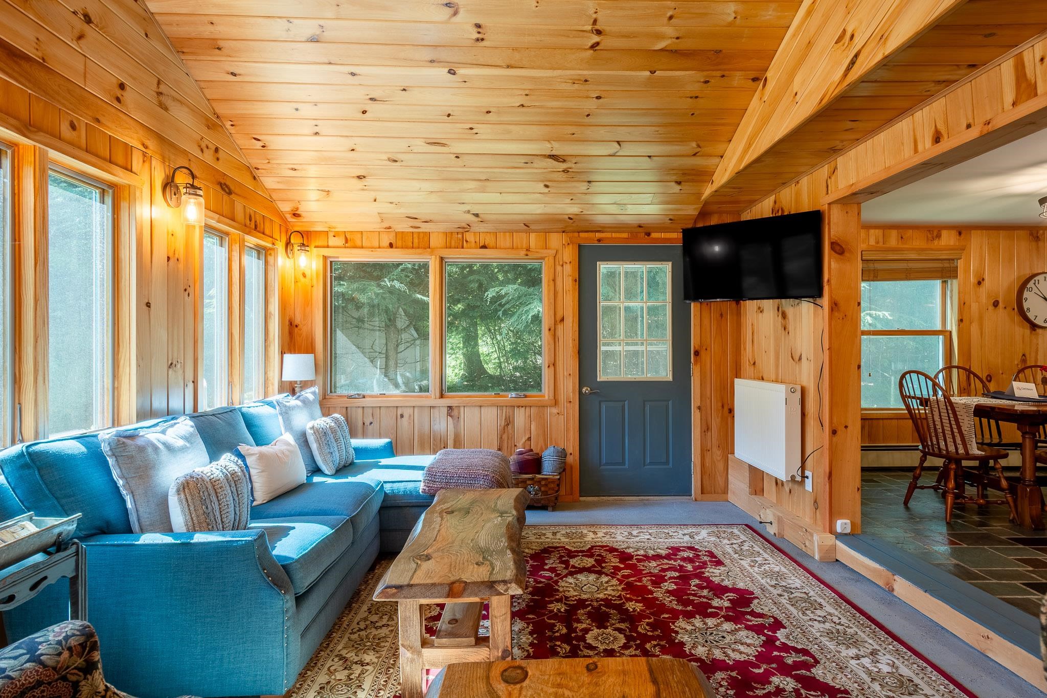 Wood-paneled living room with a blue sectional sofa and large windows overlooking trees.